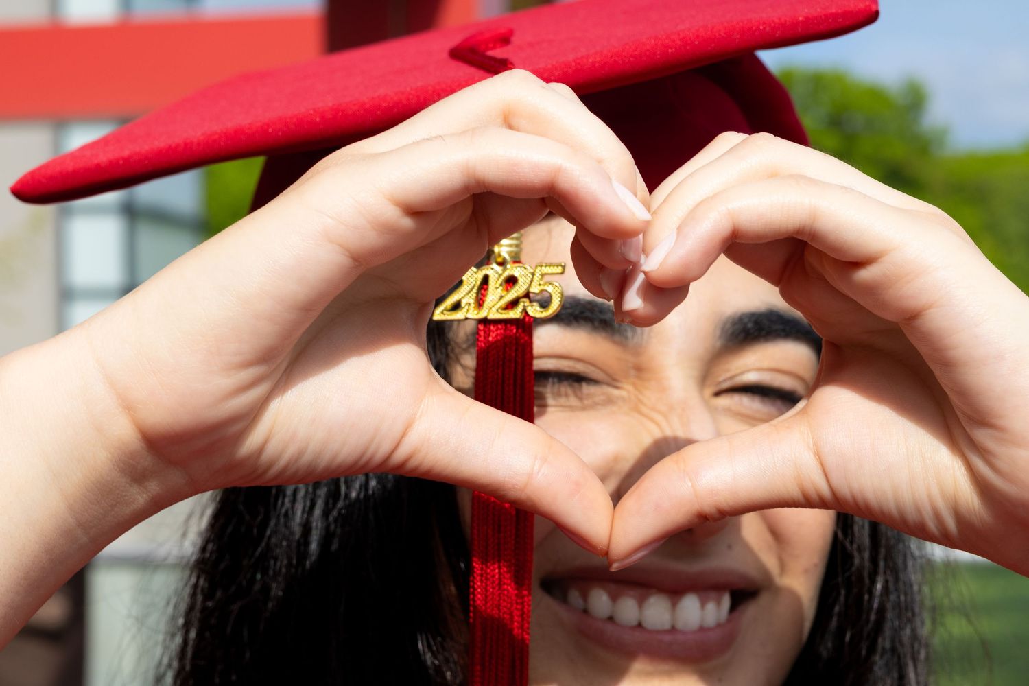 Smiling graduate making heart shape with hands showing gold 2025 charm, wearing red graduation cap outdoors.
