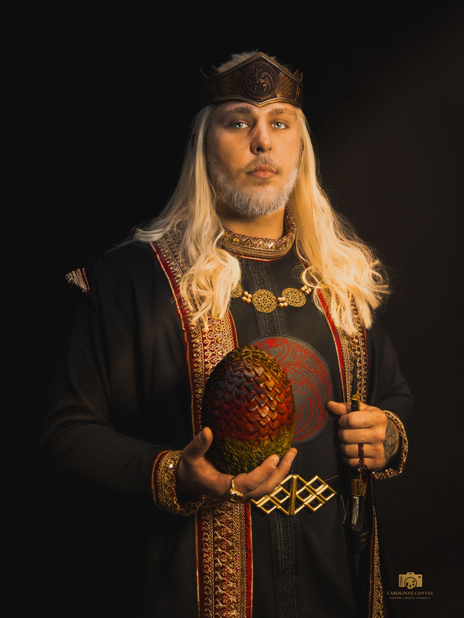 Man in medieval fantasy costume with blonde wig, crown, ornate robes, and dragon egg prop against dark background.