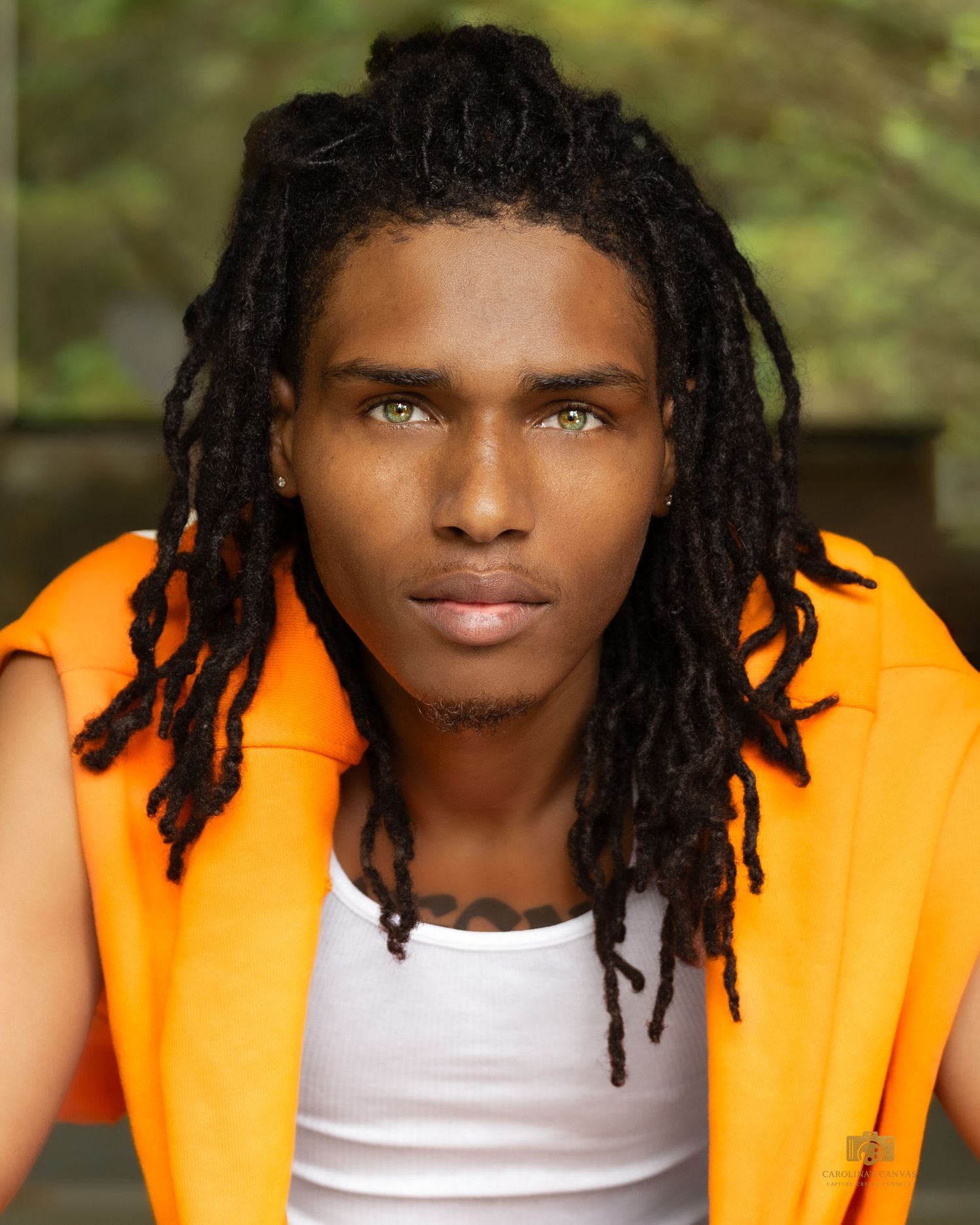 Young man with dreadlocks and green eyes wearing an orange vest over a white shirt outdoors.