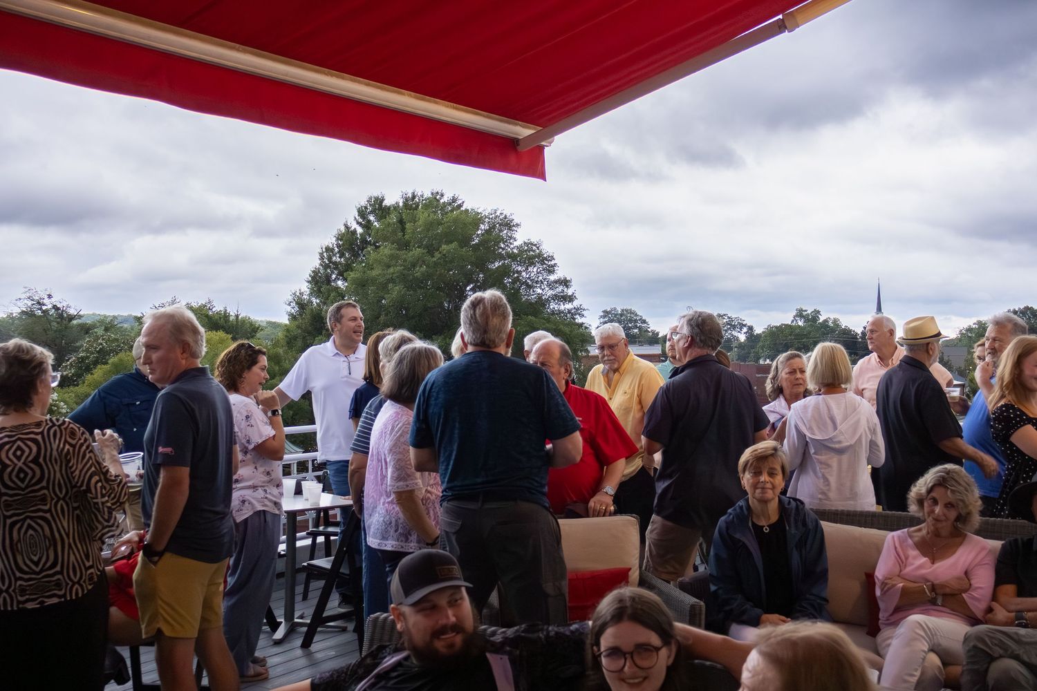 A crowded outdoor gathering of people under a red awning on a cloudy day.