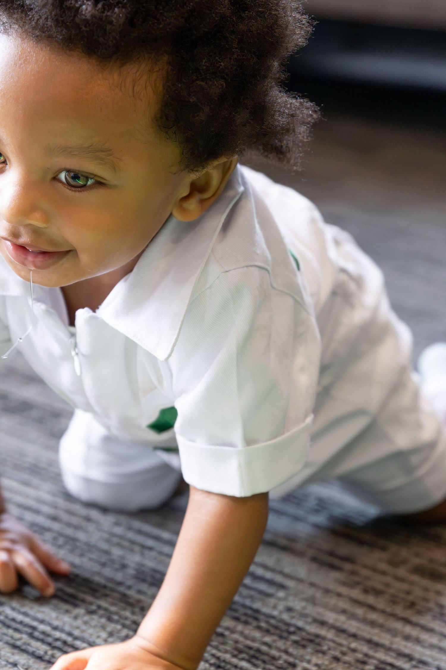 A small child in a white shirt crawls and explores on a patterned gray carpet.