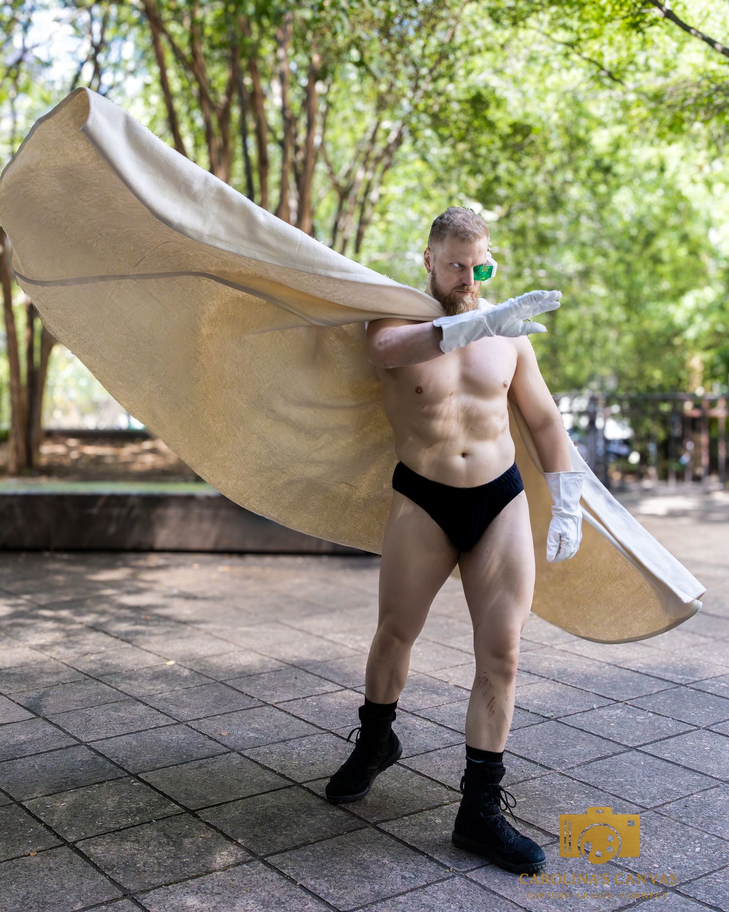 Muscular man in black briefs, white gloves, green sunglasses, and a flowing cape posing outdoors in a park.