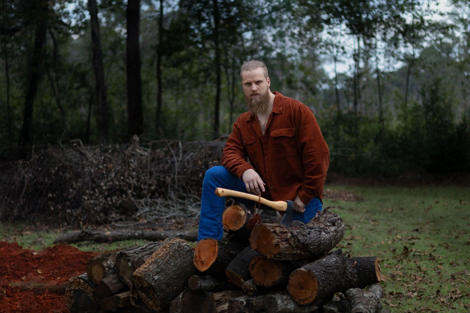 A person in a red jacket sits on stacked logs in a wooded clearing.