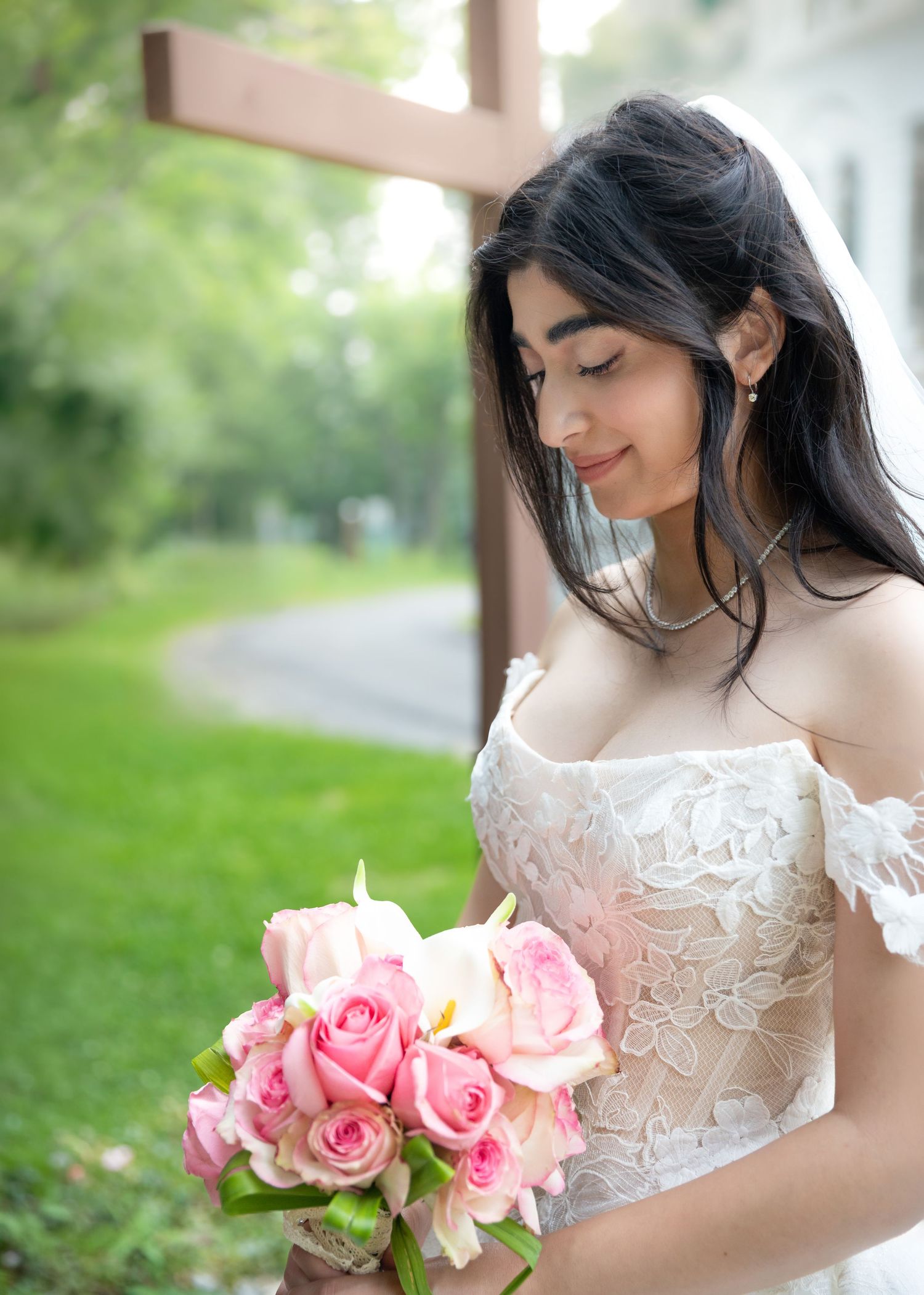 A bride in a lace off-shoulder gown holds pink roses, standing before a wooden cross outdoors.