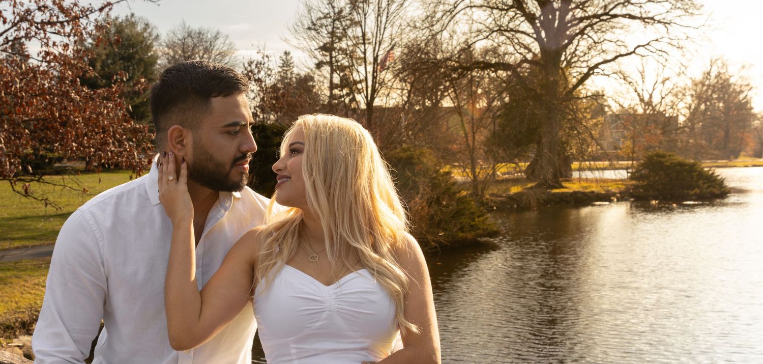 A couple in white attire shares a romantic moment by a tranquil lake during golden hour sunset.