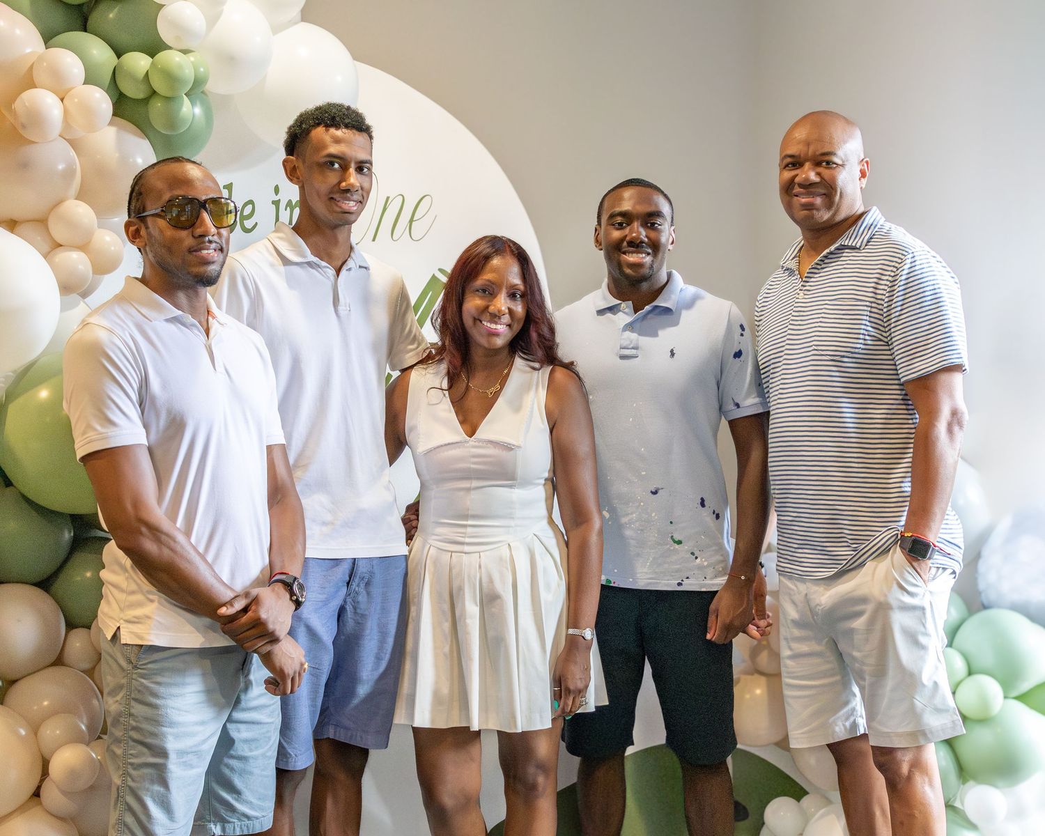 A group of people dressed in white and cream colored outfits pose together at a party with green balloon decorations.