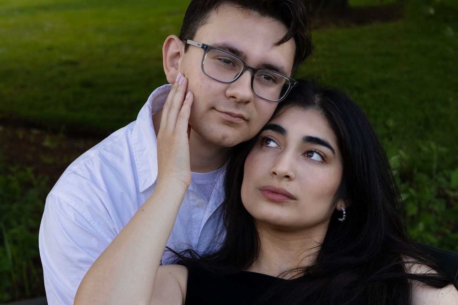 Young couple posing outdoors, man with glasses embracing woman with dark hair against green background.