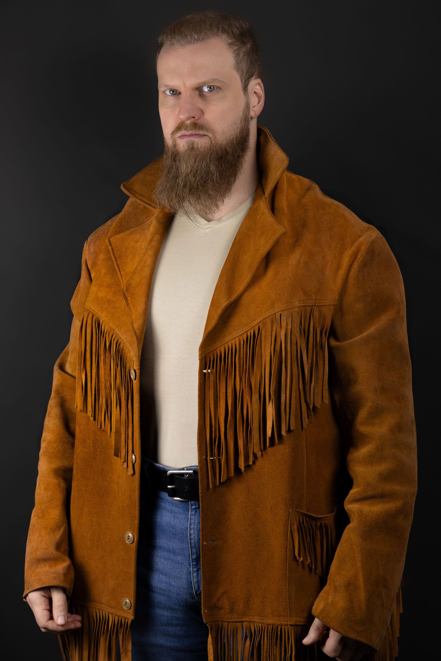 Person wearing a suede western-style fringe jacket in brown with a cream colored shirt and blue jeans against dark background.