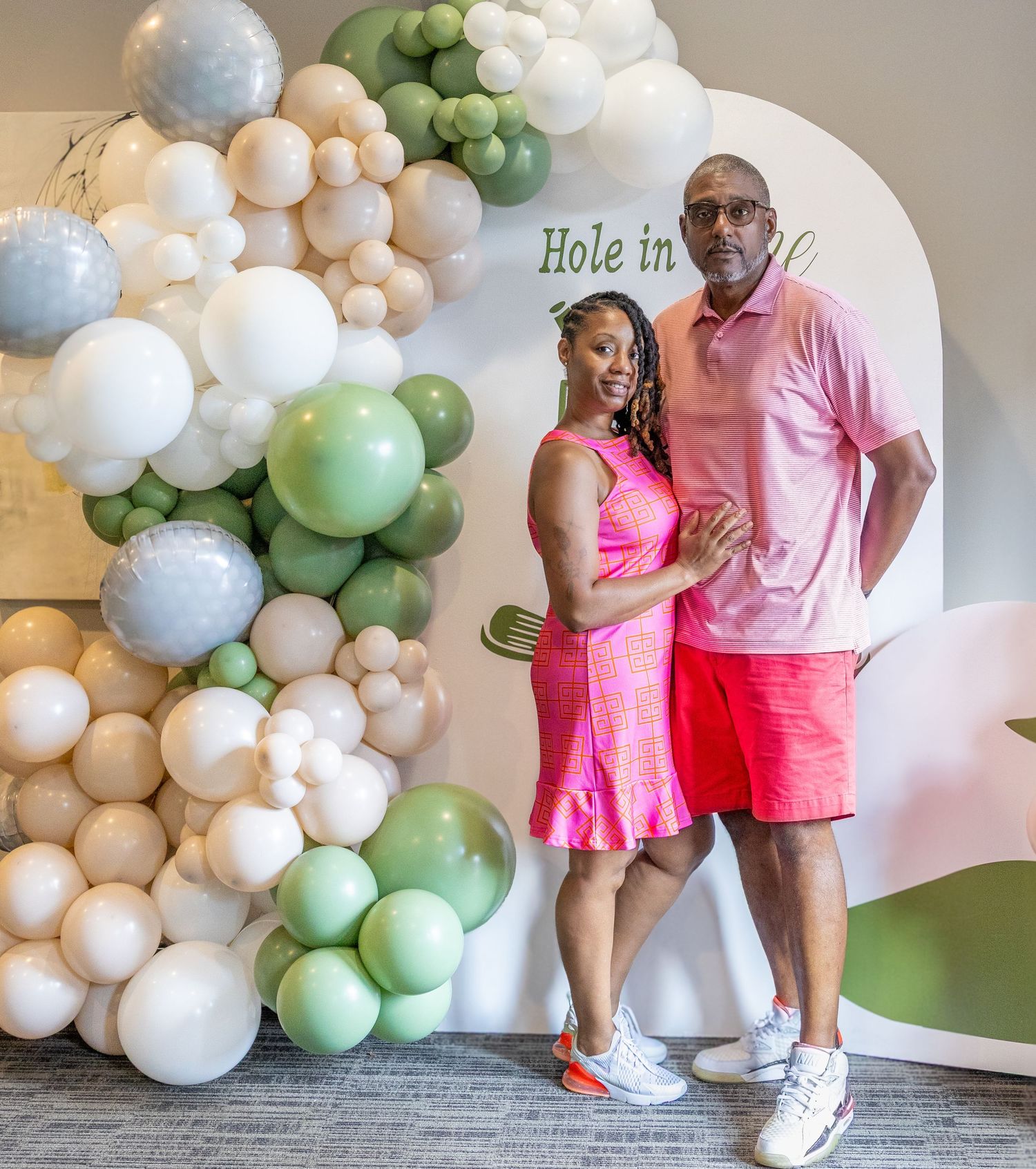 A couple poses together in matching pink outfits against a decorative balloon arch with white and sage green colors.