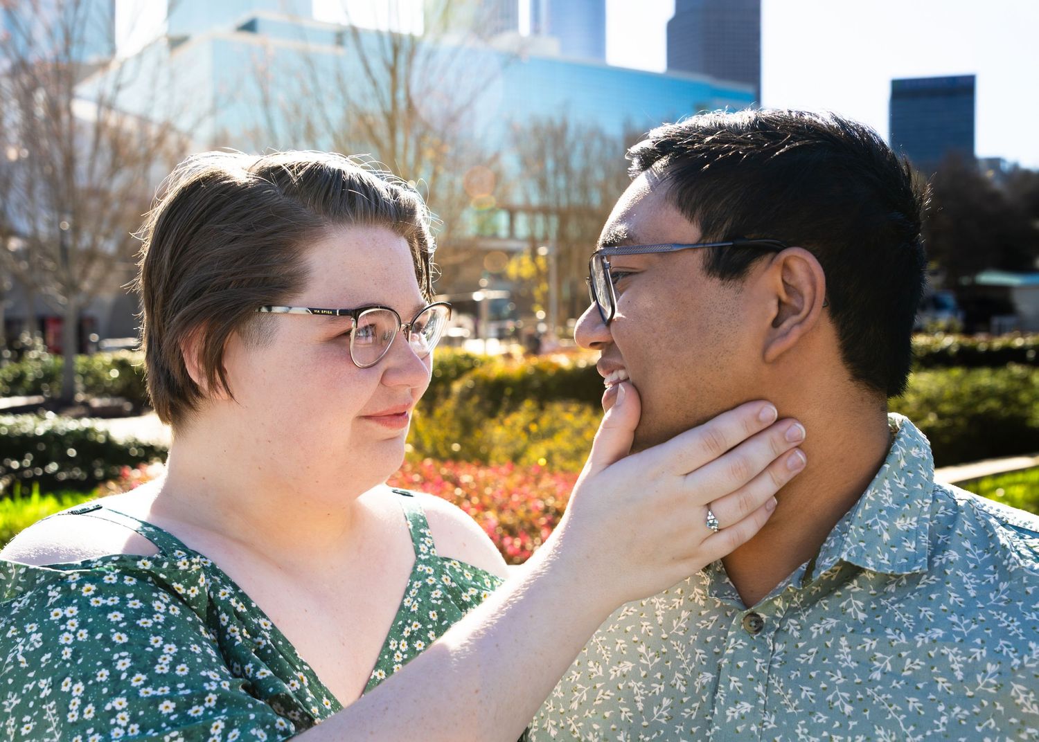 Couple sharing a tender moment outdoors, woman in floral dress touching mans face, city park background.