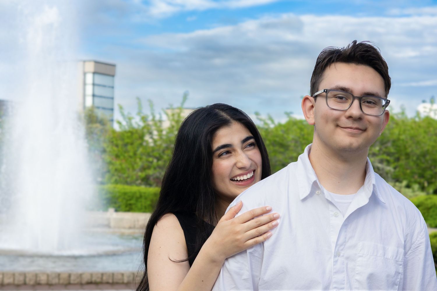 A smiling couple poses near a decorative fountain outdoors, with lush green trees and a building in the background.