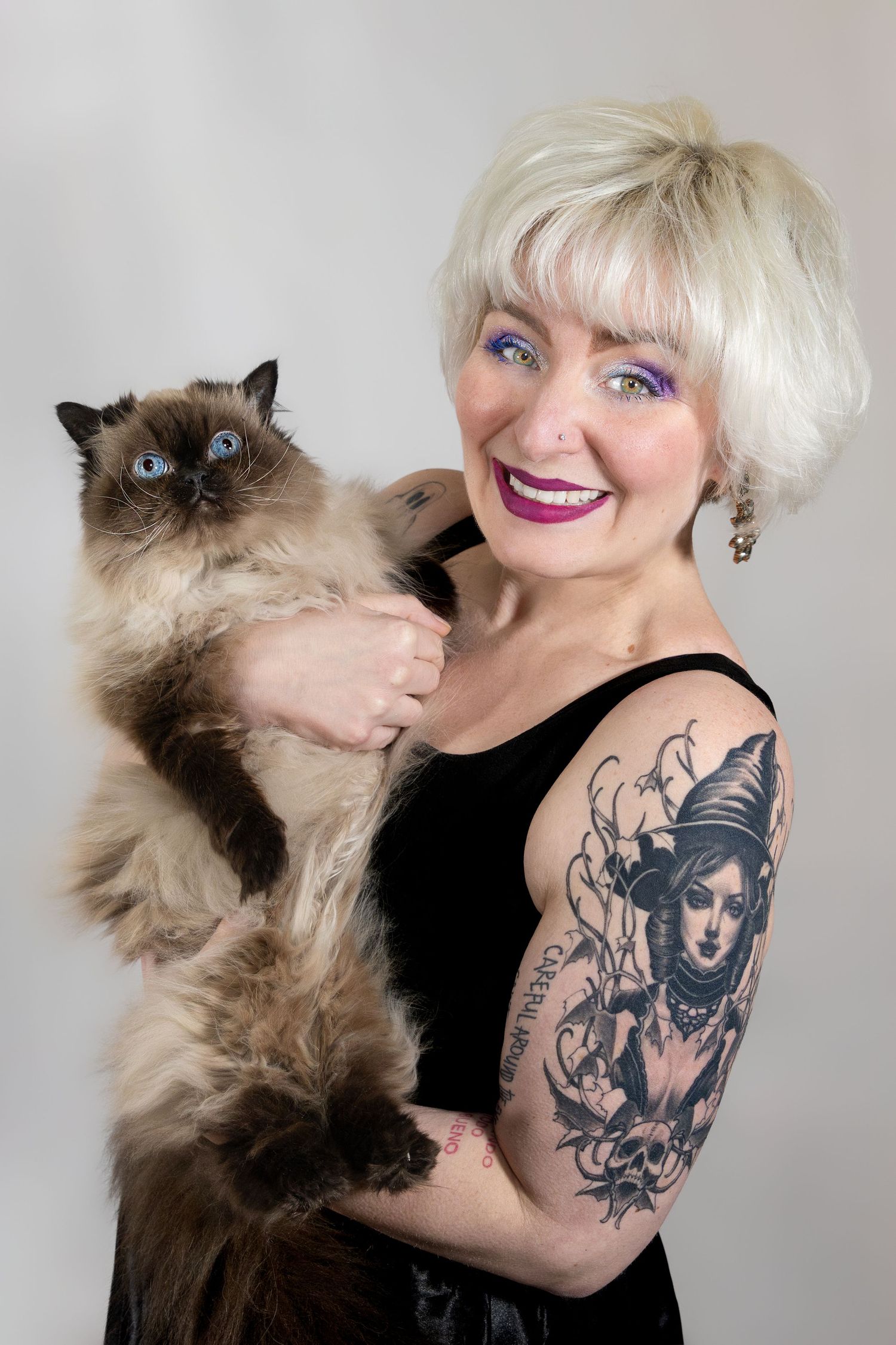 Smiling blonde woman with tattoo sleeve holding a fluffy Himalayan cat with blue eyes against gray background.