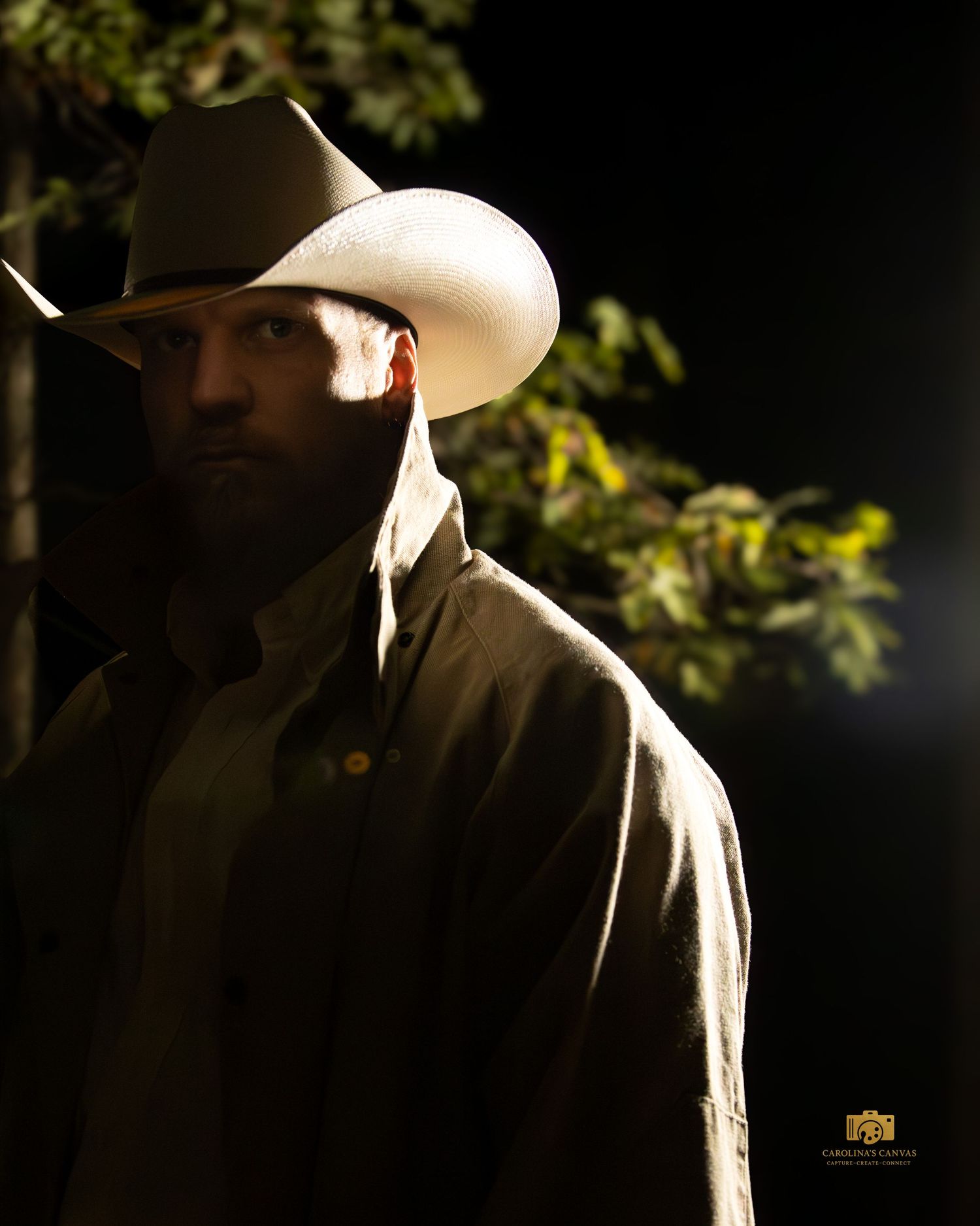A figure in a cowboy hat stands in dramatic lighting against a dark background with foliage visible.