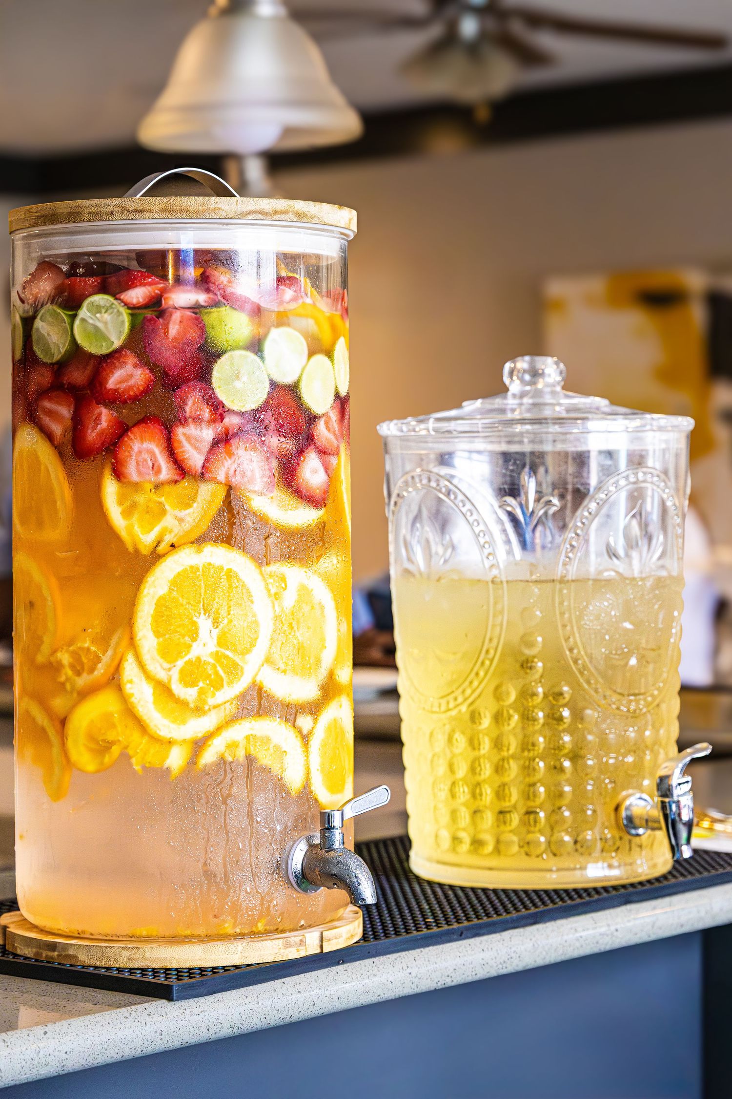 Glass drink dispensers filled with infused water featuring fresh citrus slices and berries arranged in a line on a bar counter.
