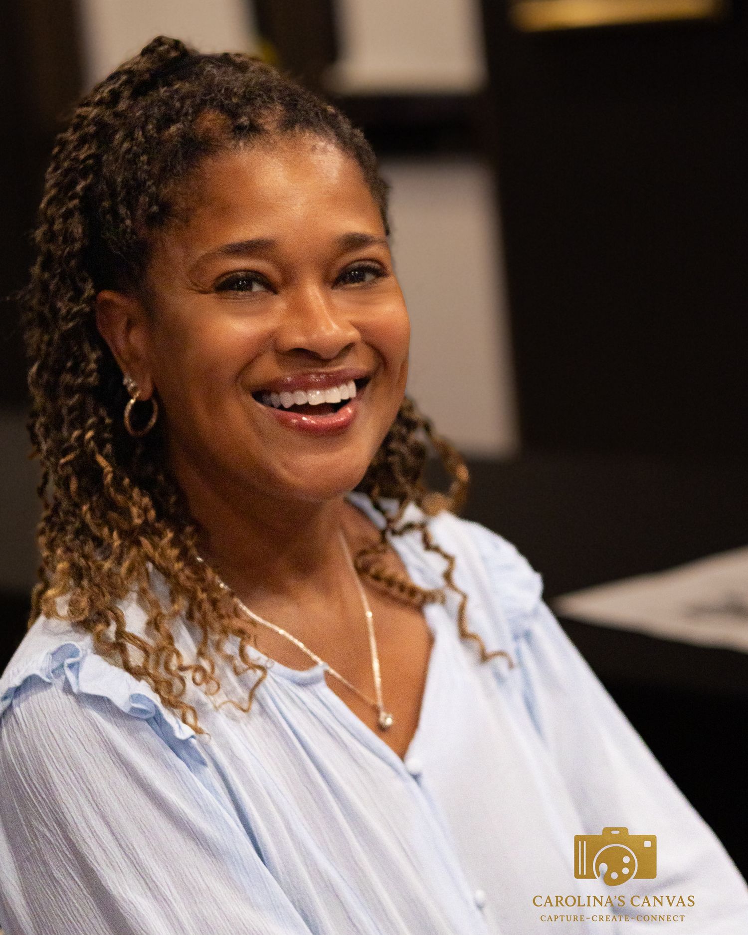 A person with curly ombre hair and silver hoop earrings smiles warmly while wearing a light blue button-down shirt.