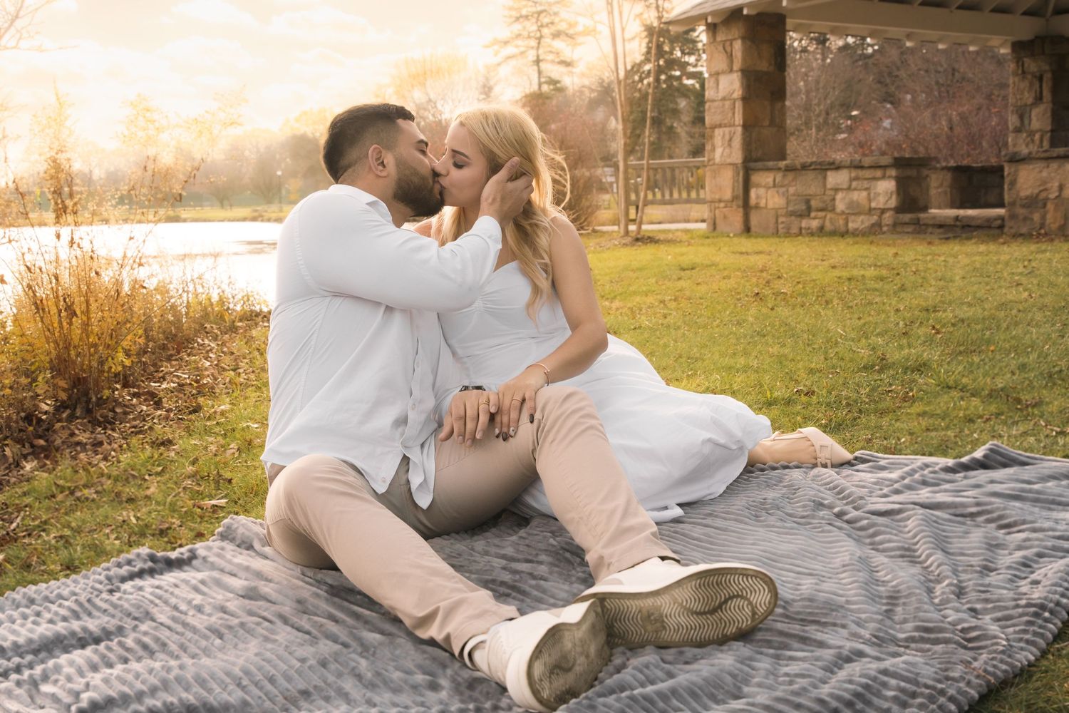 A couple shares a romantic kiss on a picnic blanket in a sunlit park during golden hour.