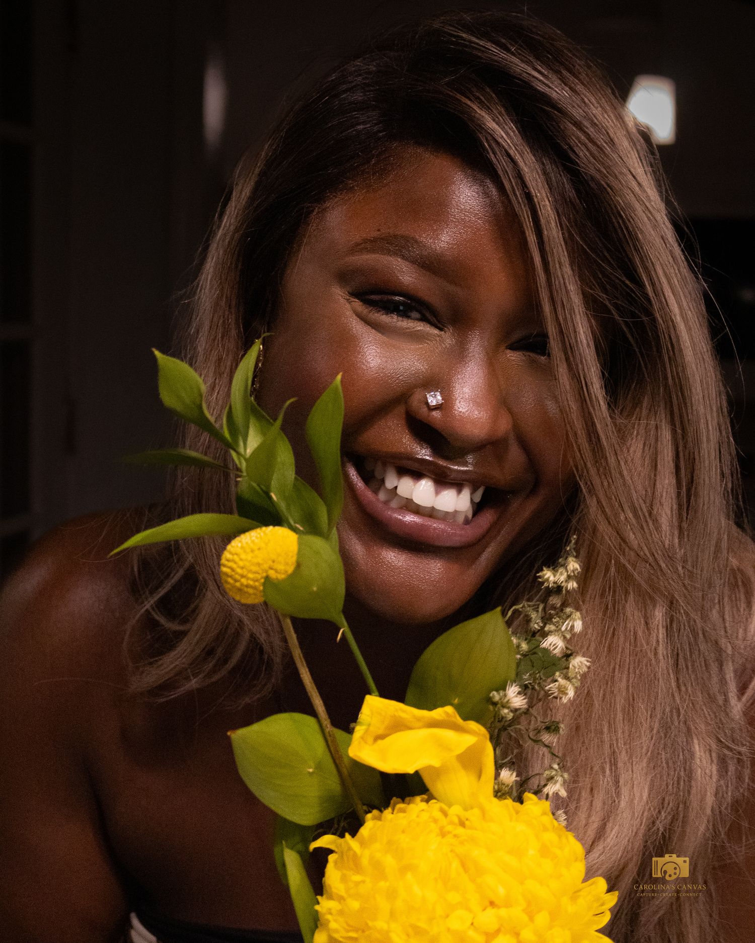 Smiling young woman with nose piercing holding vibrant yellow flowers and greenery.