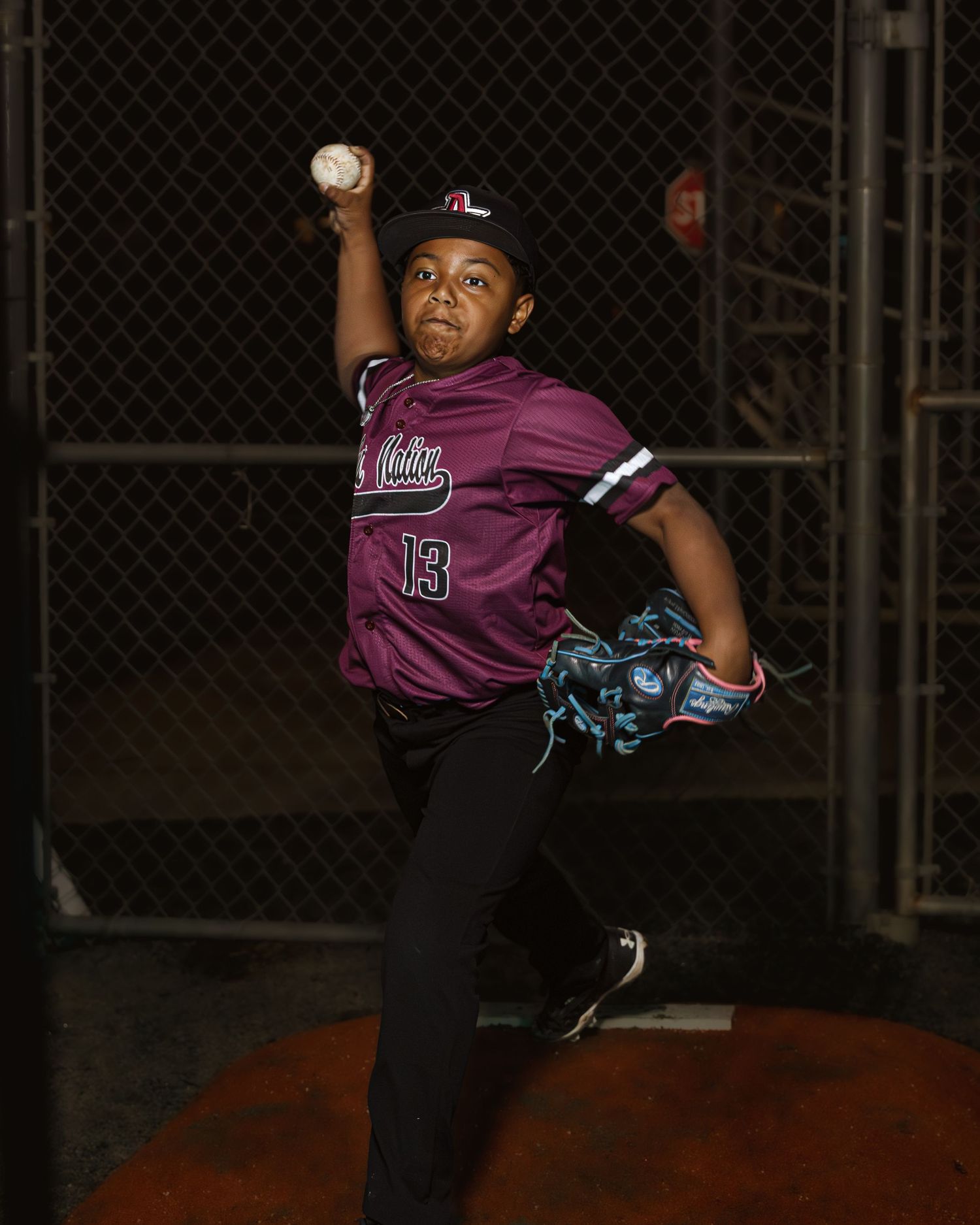 Baseball player in purple jersey mid-pitch against chain link fence at night.