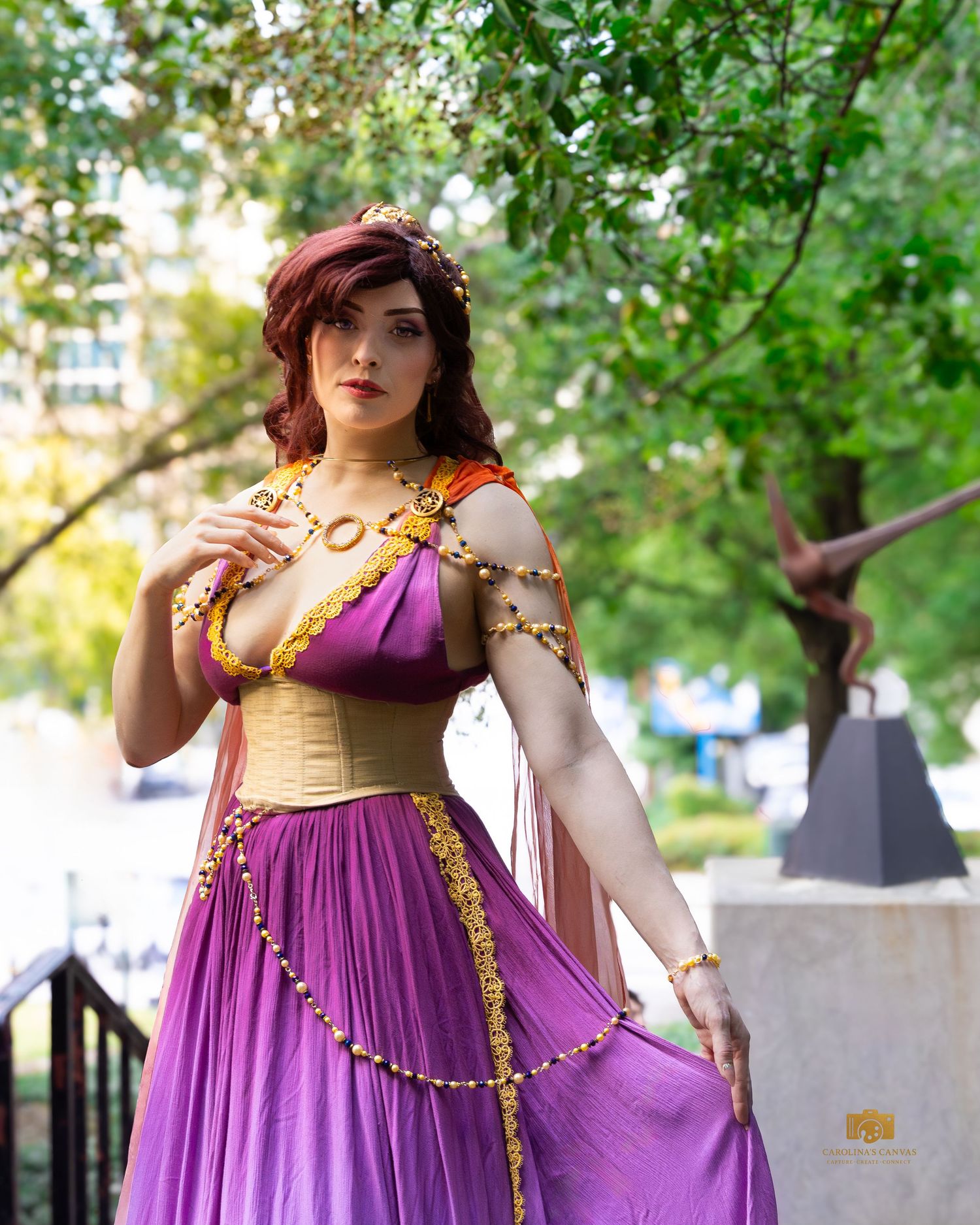 Woman in purple Grecian costume with gold accessories and red hair poses outdoors at a cosplay event.