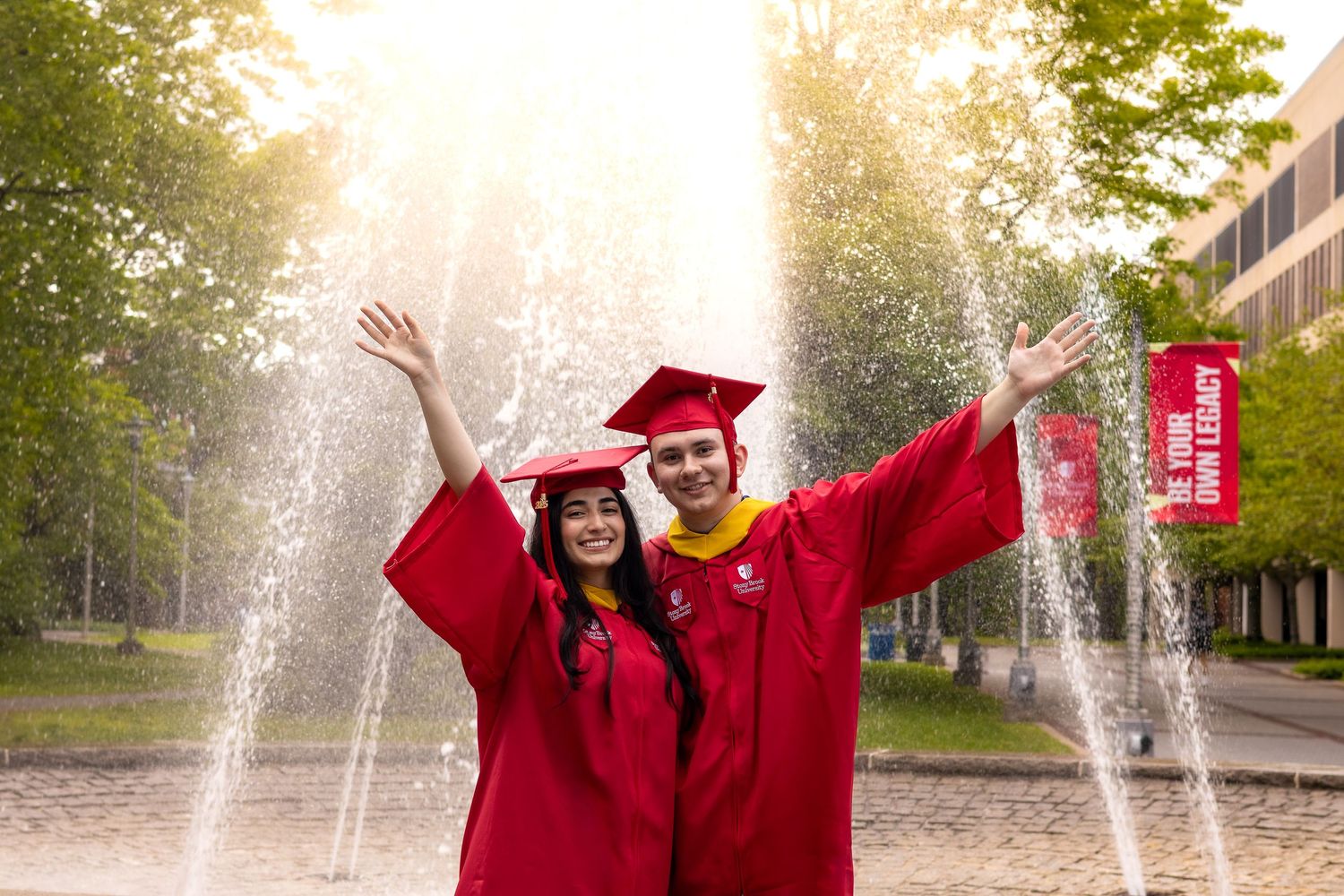 Two graduates in red caps and gowns celebrate joyfully in front of a fountain on a sunny campus day.