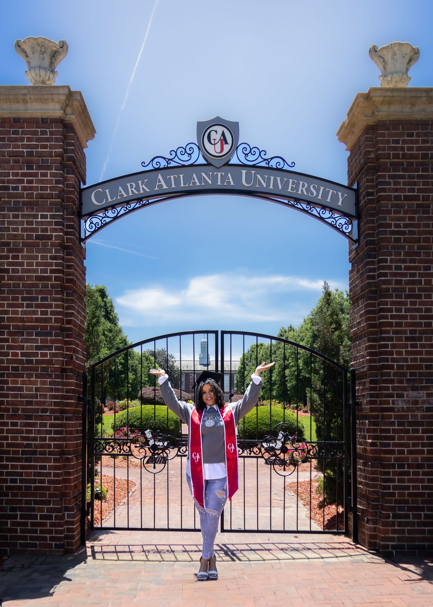 A graduate poses triumphantly in front of the iconic brick entrance gate at Clark Atlanta University on a sunny day.