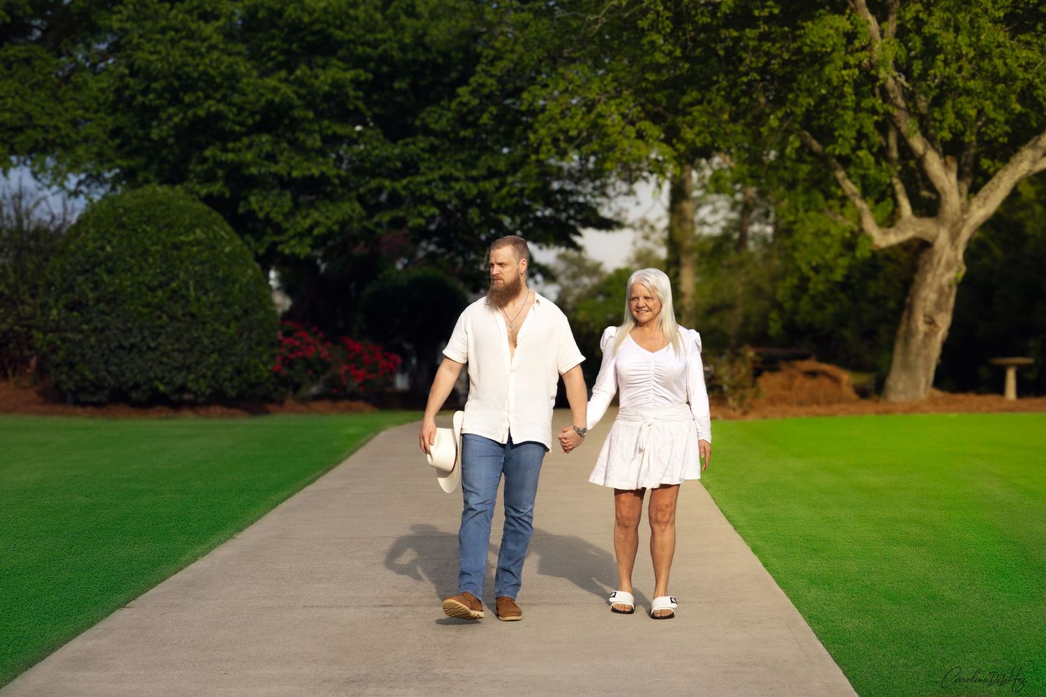 A mother and son walks hand in hand along a paved walkway through a lush green lawn surrounded by trees.