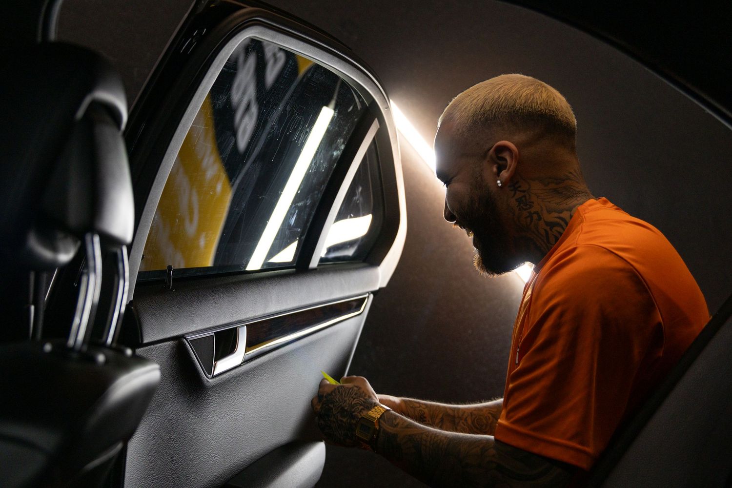 Professional detailer in orange shirt carefully inspects and works on a car door panel in dim lighting.