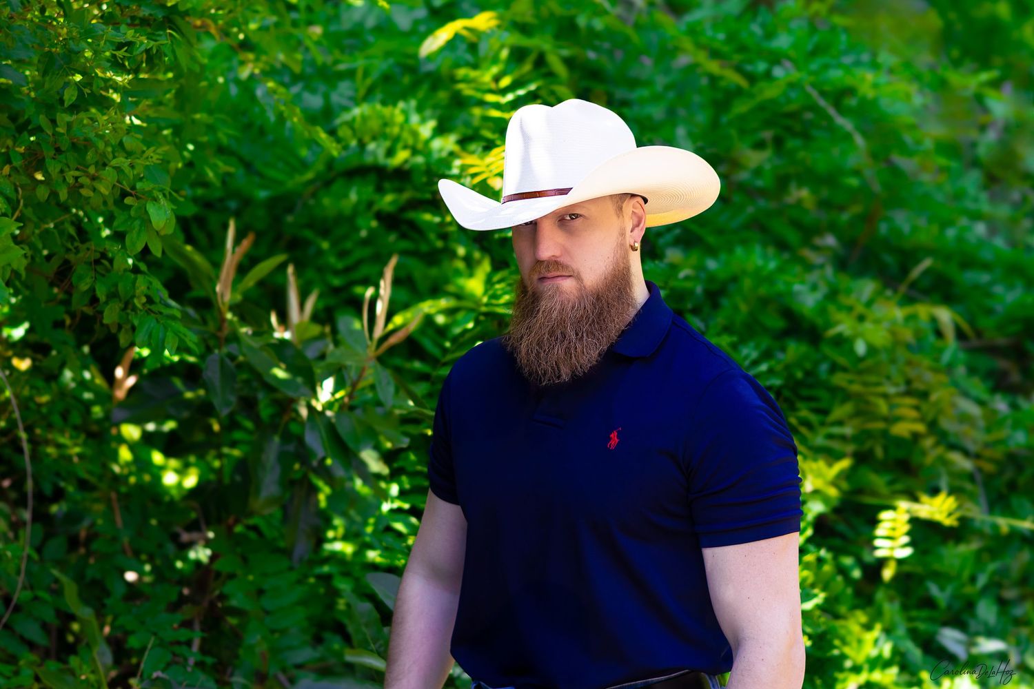 Bearded person in a white cowboy hat and navy polo shirt stands against lush green foliage outdoors.