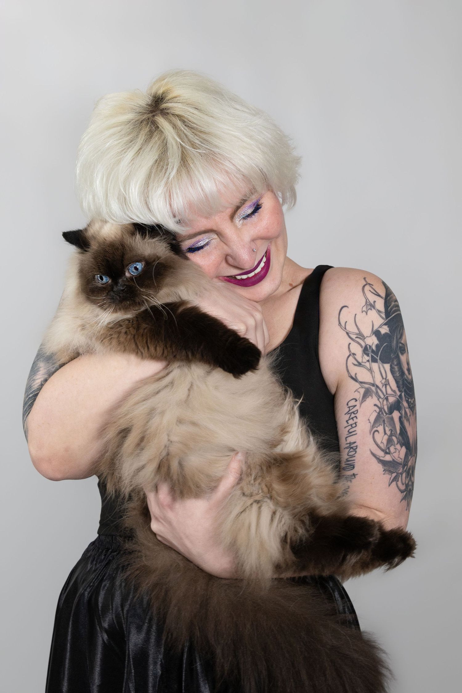 Tattooed blonde woman with pink makeup smiling while holding a fluffy Siamese cat in a black dress.