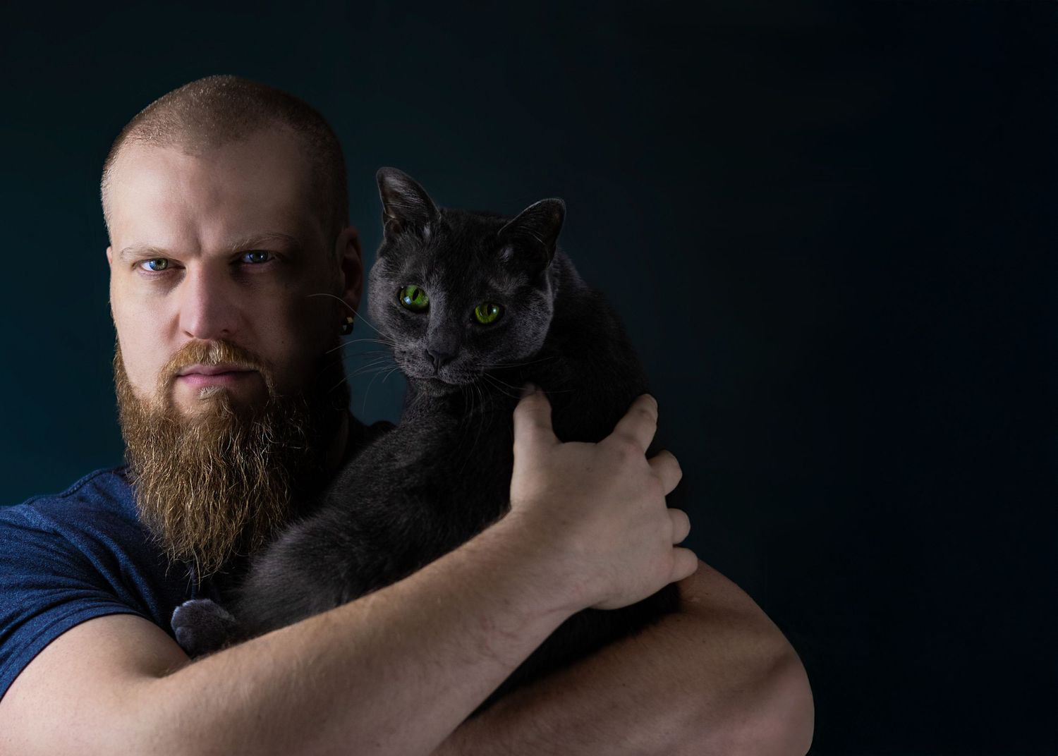 A bearded man in a dark shirt holds a black cat against a dark background for a portrait.
