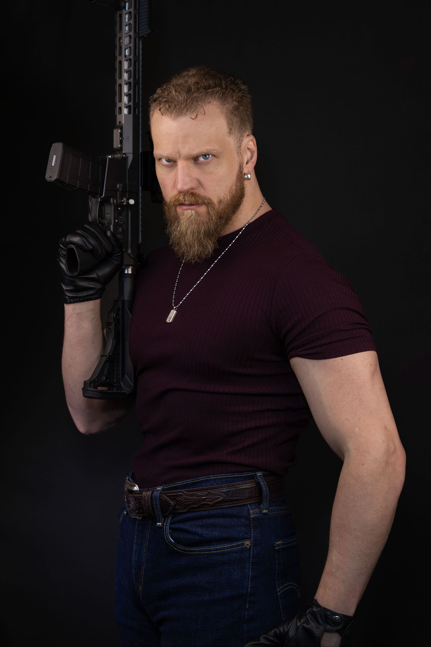 Portrait against dark background wearing burgundy t-shirt and jeans showing muscular arms and beard. Headshots