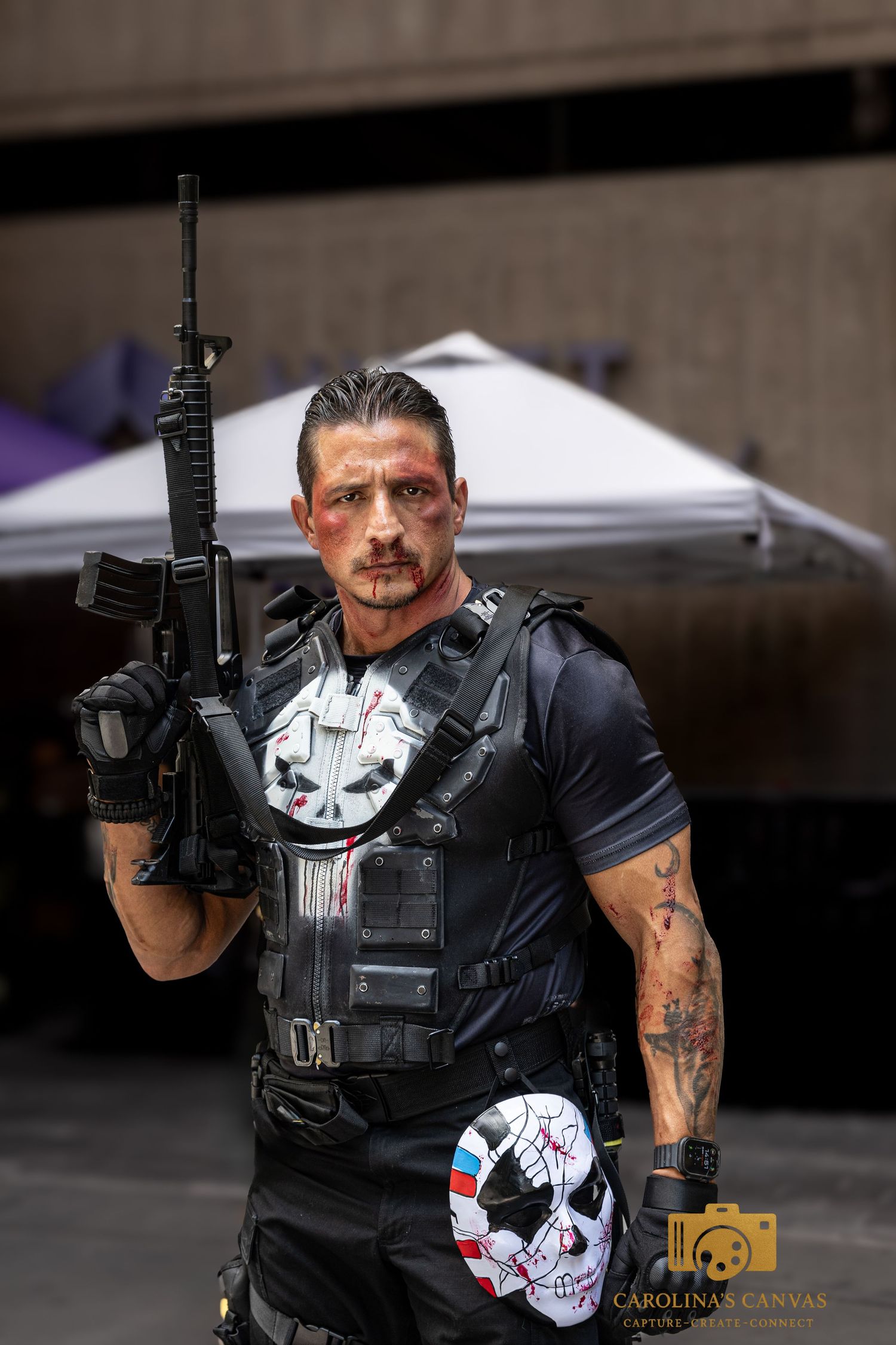 A cosplayer wearing tactical gear and a skull mask poses with an assault rifle prop against a dark background.