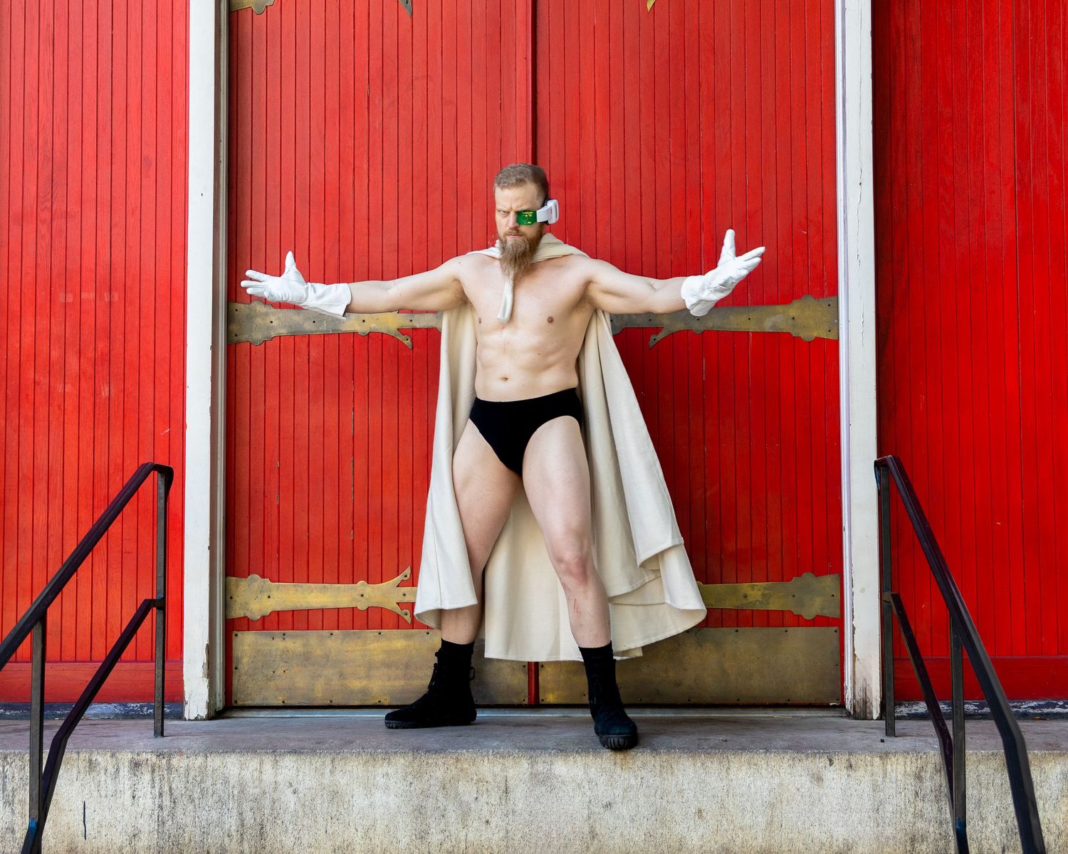 A figure in a beige coat poses dramatically against a bright red wall with corrugated metal siding.
