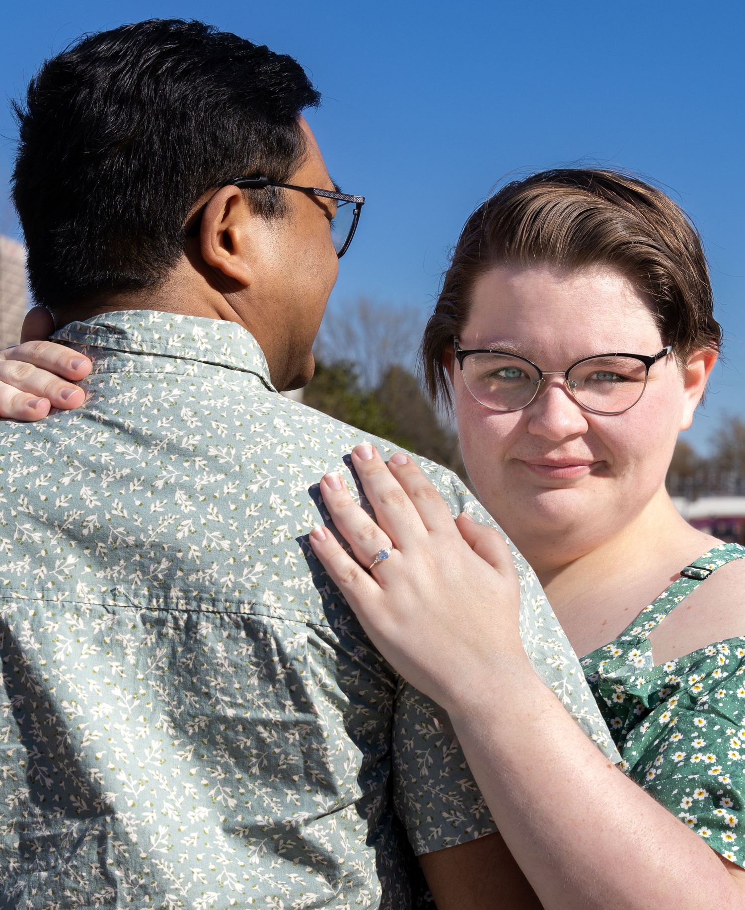 A couple in floral shirts embrace outdoors on a sunny day, woman with short hair and glasses smiling warmly.