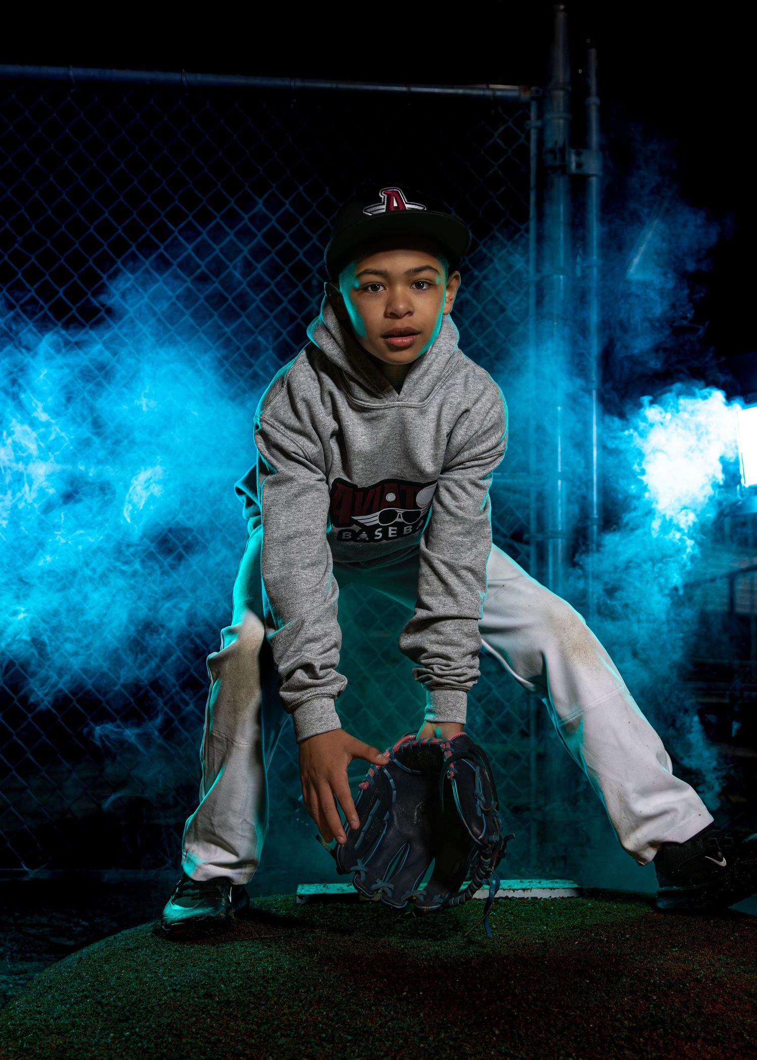 Baseball player in grey hoodie crouching with glove against blue smoke effect.