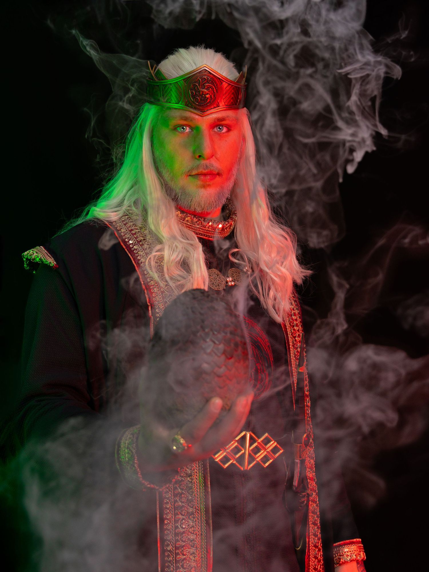 Fantasy king cosplayer with white hair, red crown, dark robes, holding a dragon egg amid swirling smoke with green lighting.