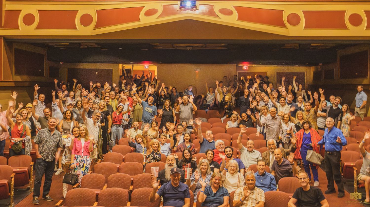 A large audience fills theater seats with orange chairs while standing and raising their hands in excitement.
