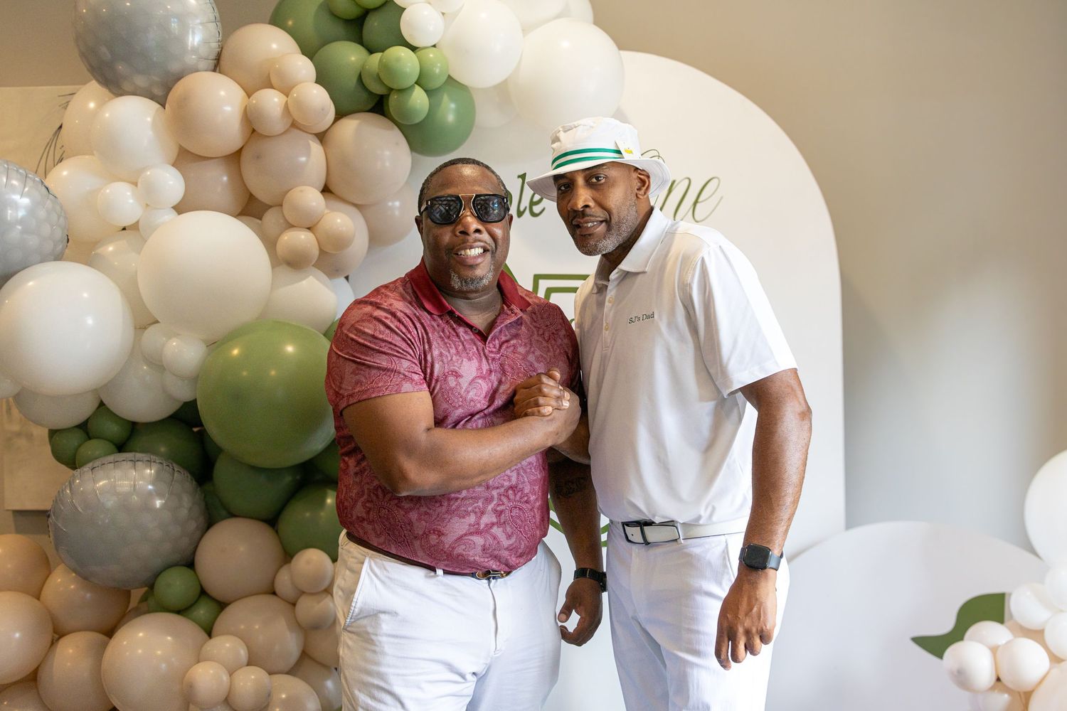 Two people pose together in front of a decorative balloon arch with white, green and silver balloons.