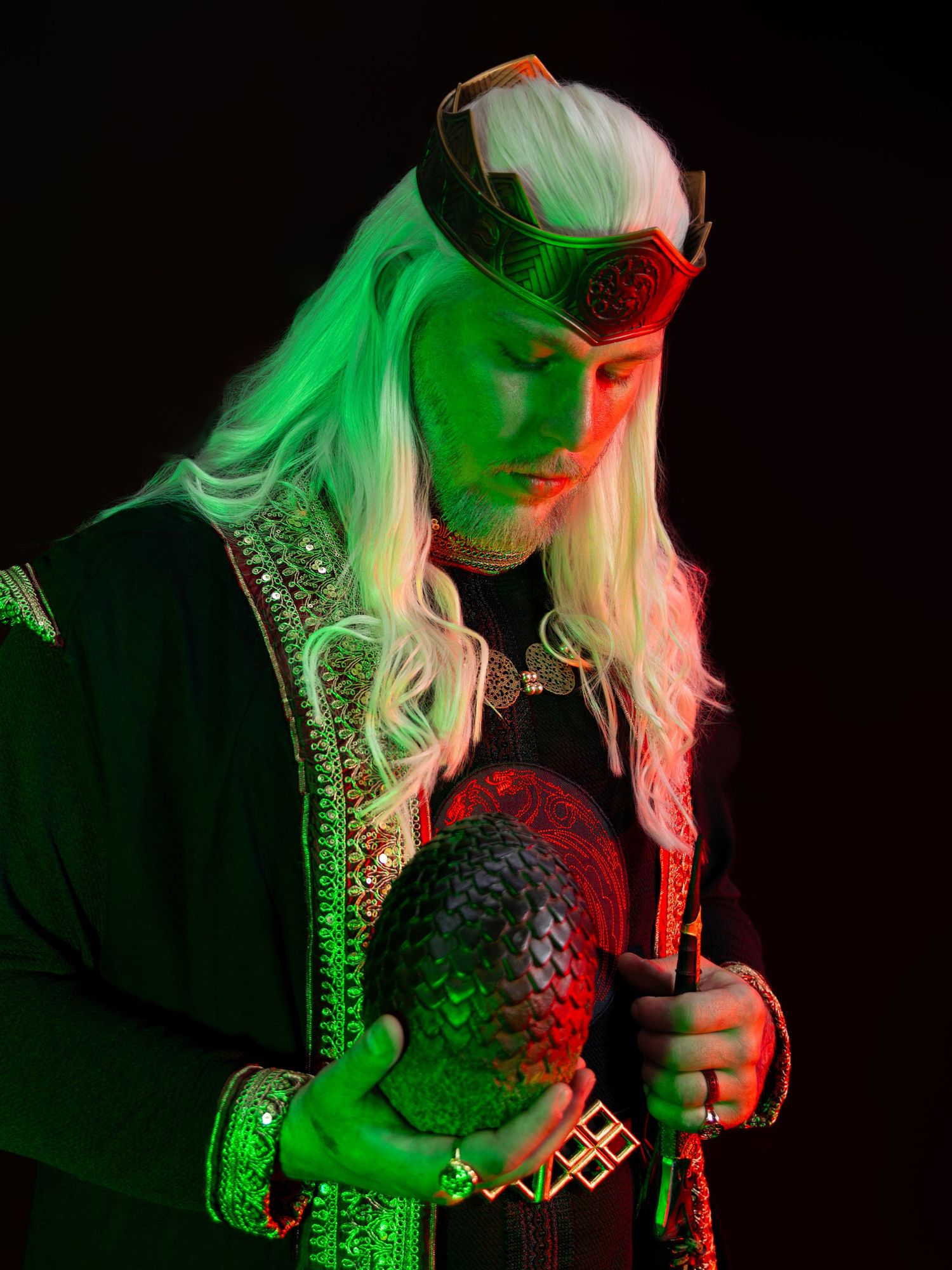 Person in fantasy costume with white wig, crown, and dragon egg under dramatic green and red lighting.