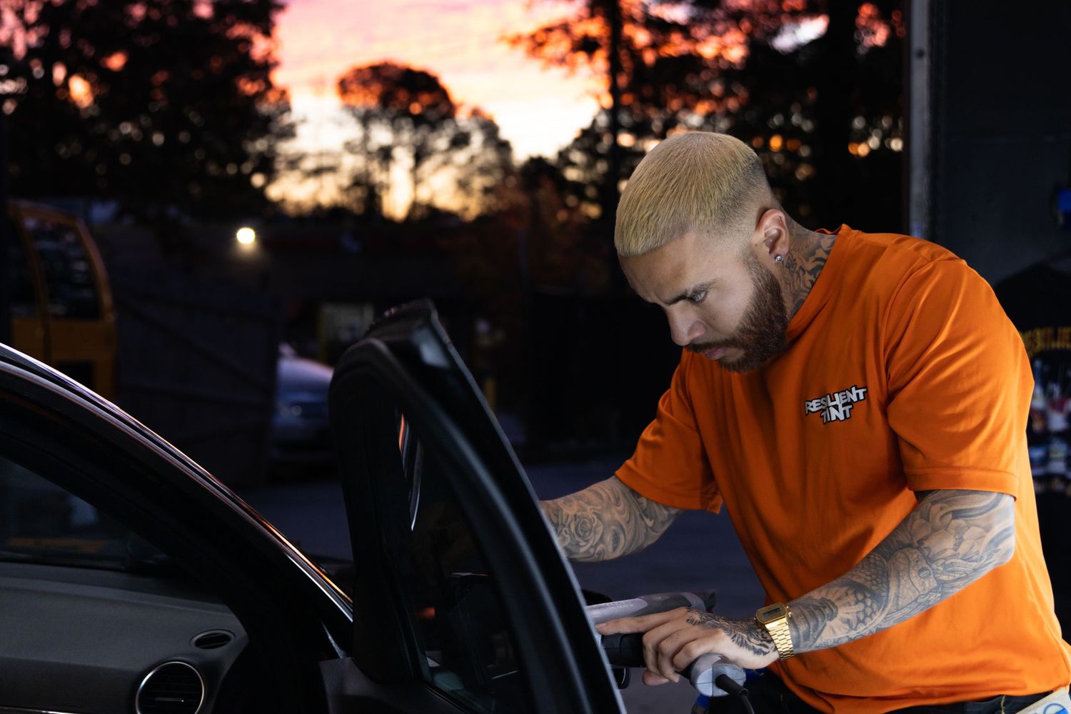 Auto detailing technician in orange uniform works on a car exterior during sunset with trees in background.