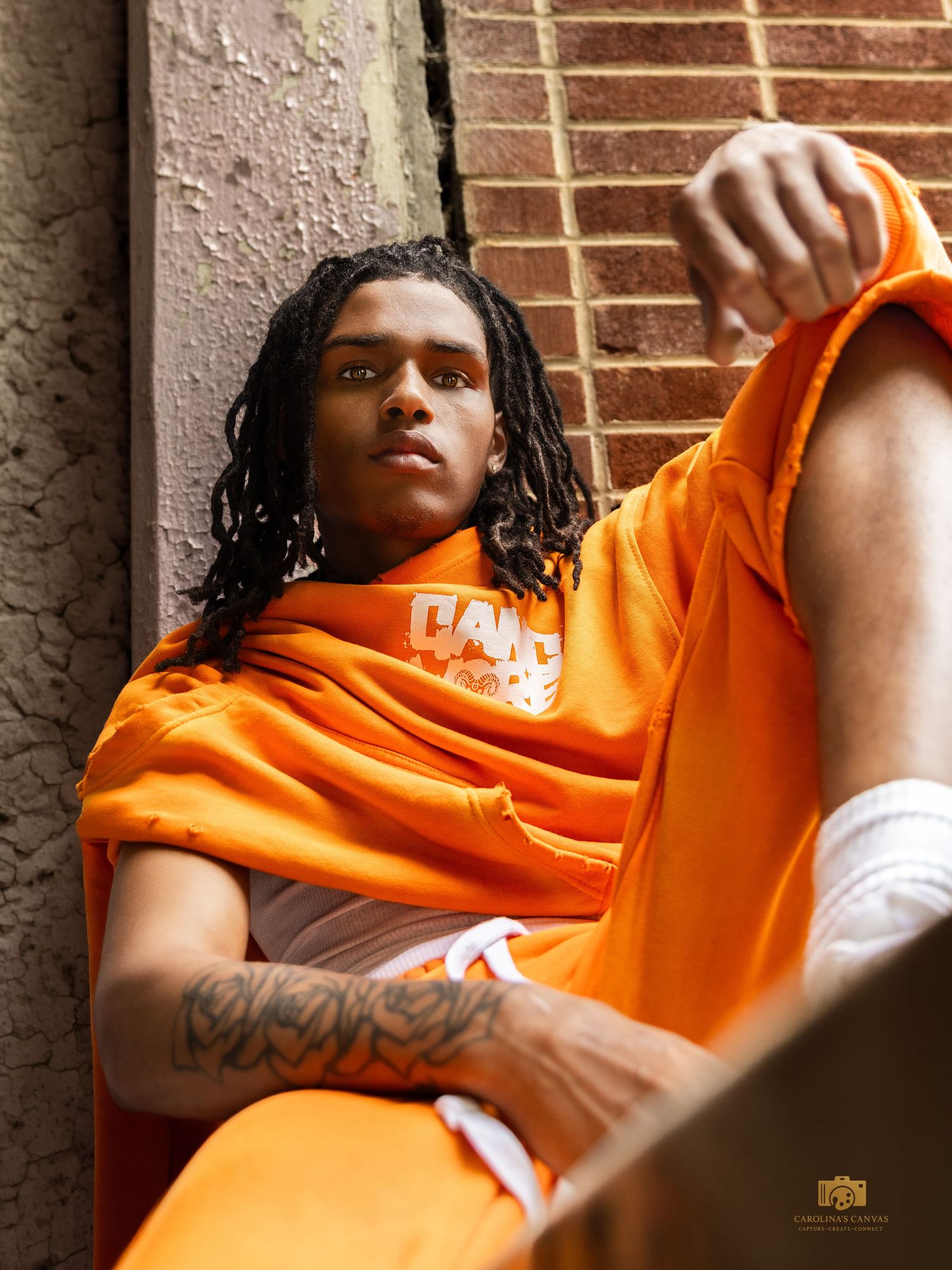 Young man with dreadlocks in orange hoodie and shorts leaning against brick wall with tattoos visible.