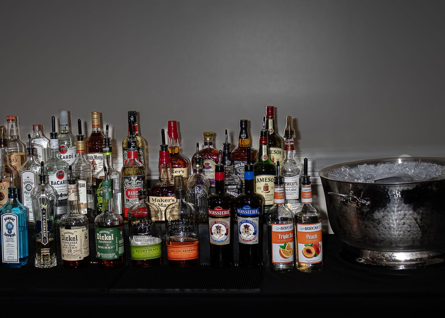 Silhouetted liquor bottles line up against a dark background creating a moody bar atmosphere.