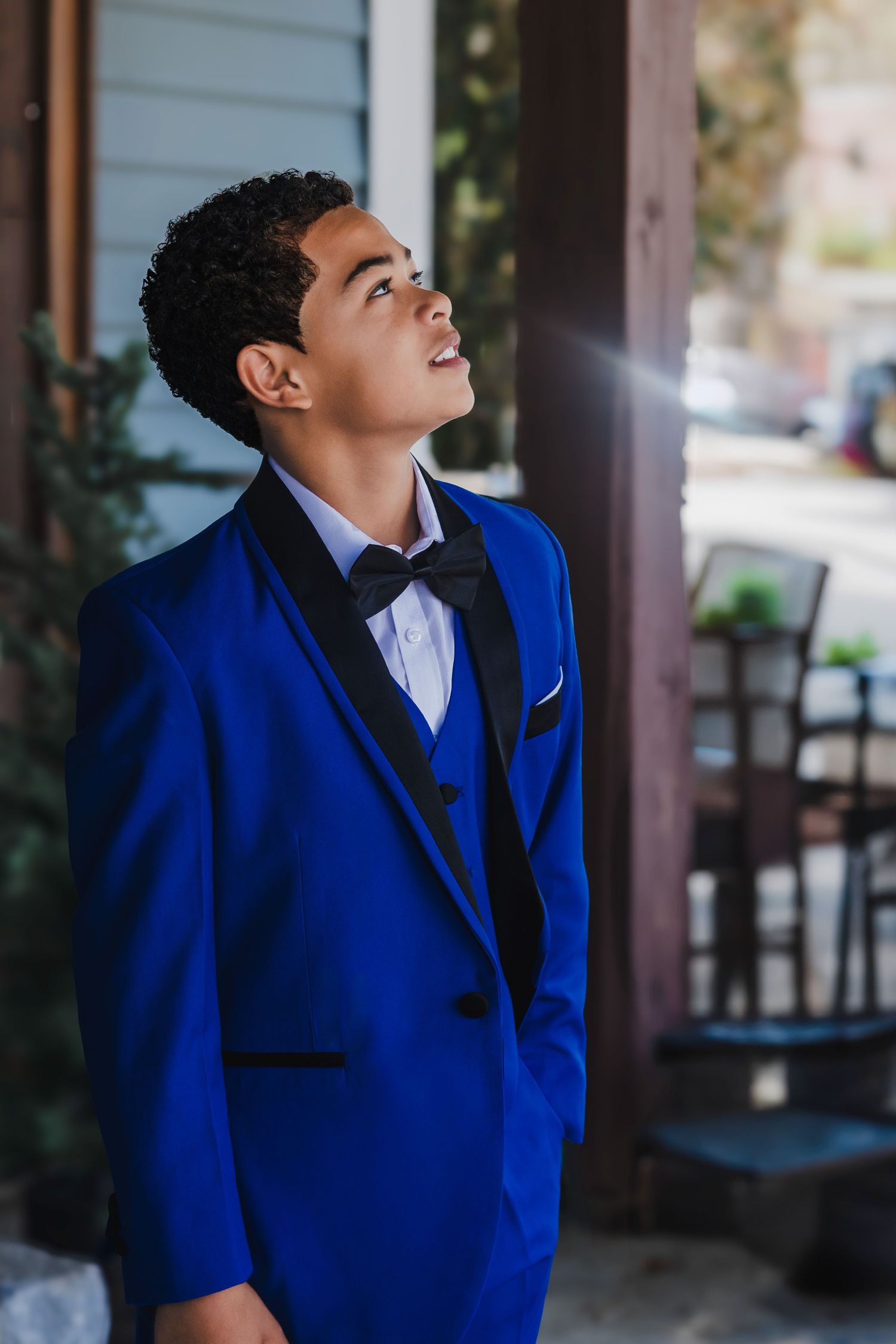 A young person in a royal blue tuxedo with black bow tie looks upward with a bright smile.