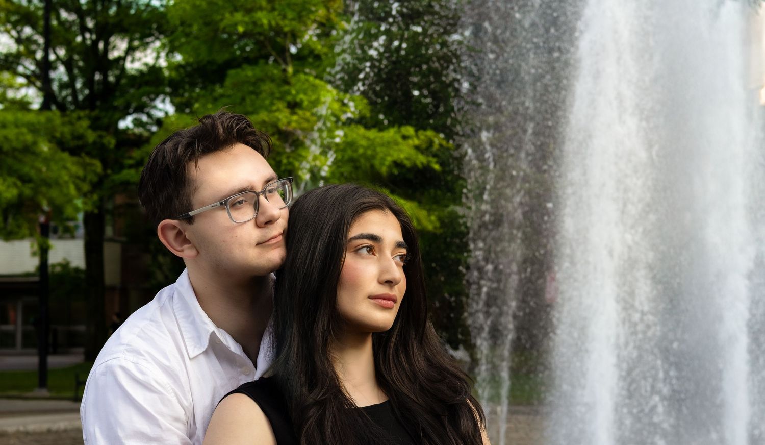 Young couple posing together near a decorative fountain outdoors, surrounded by lush green trees.