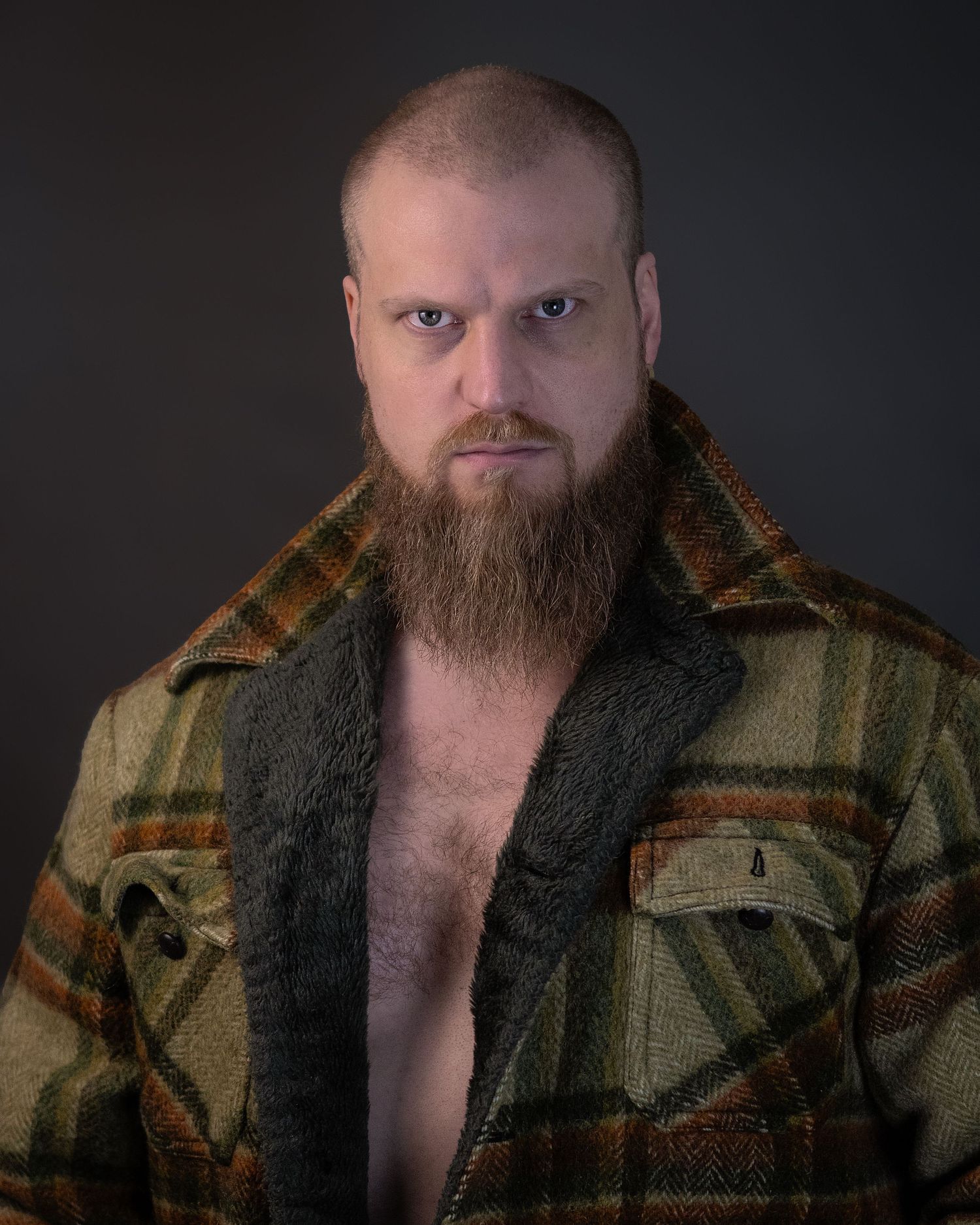 Portrait of a bearded person in a plaid flannel jacket against dark background.