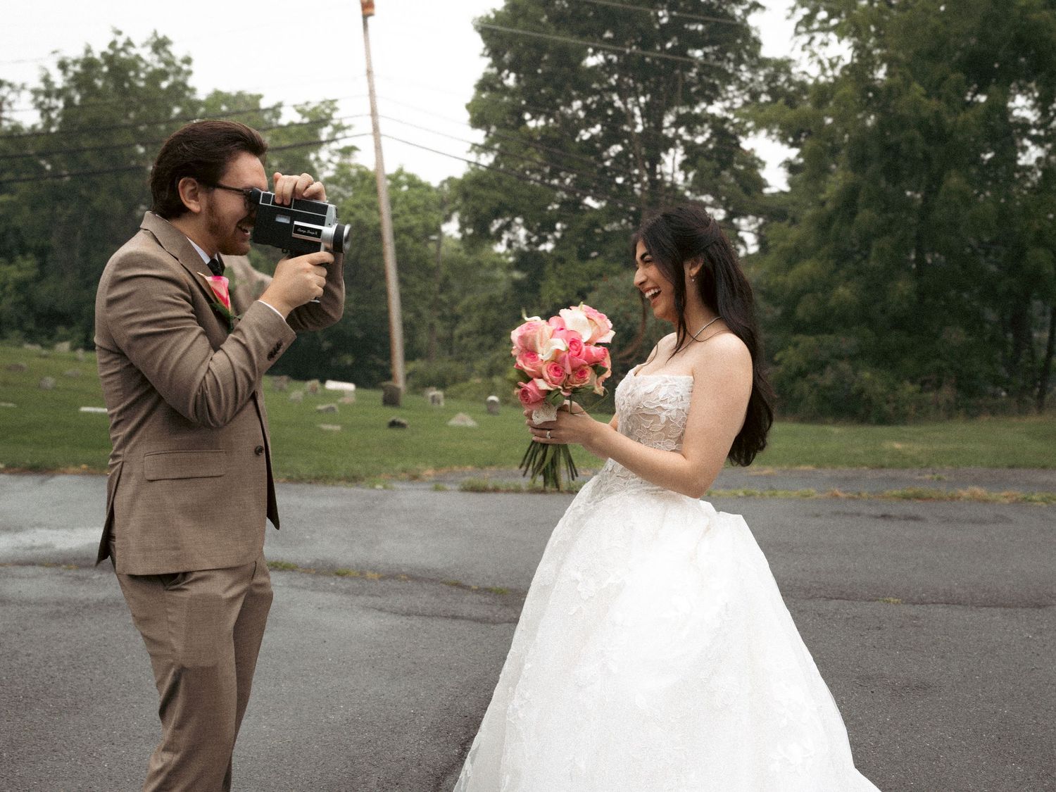 Groom in tan suit photographs bride in white gown holding pink bouquet outdoors on wedding day.