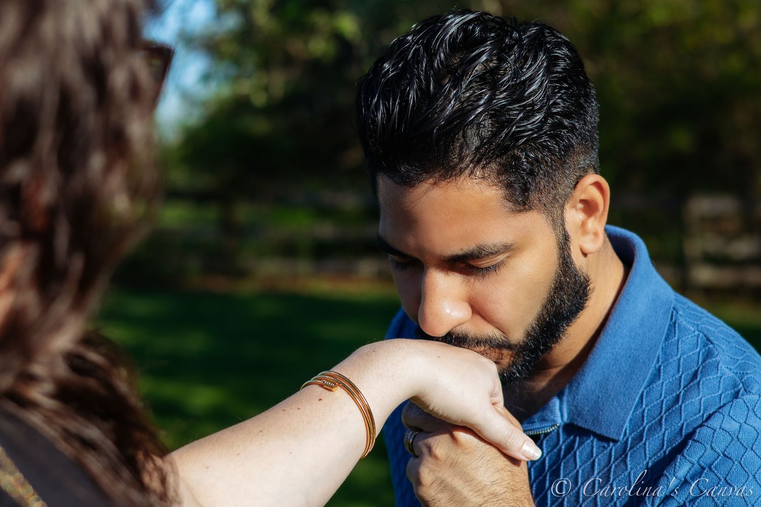 A bearded man in a blue button-down shirt gently kisses a person's hand outdoors on a sunny day.