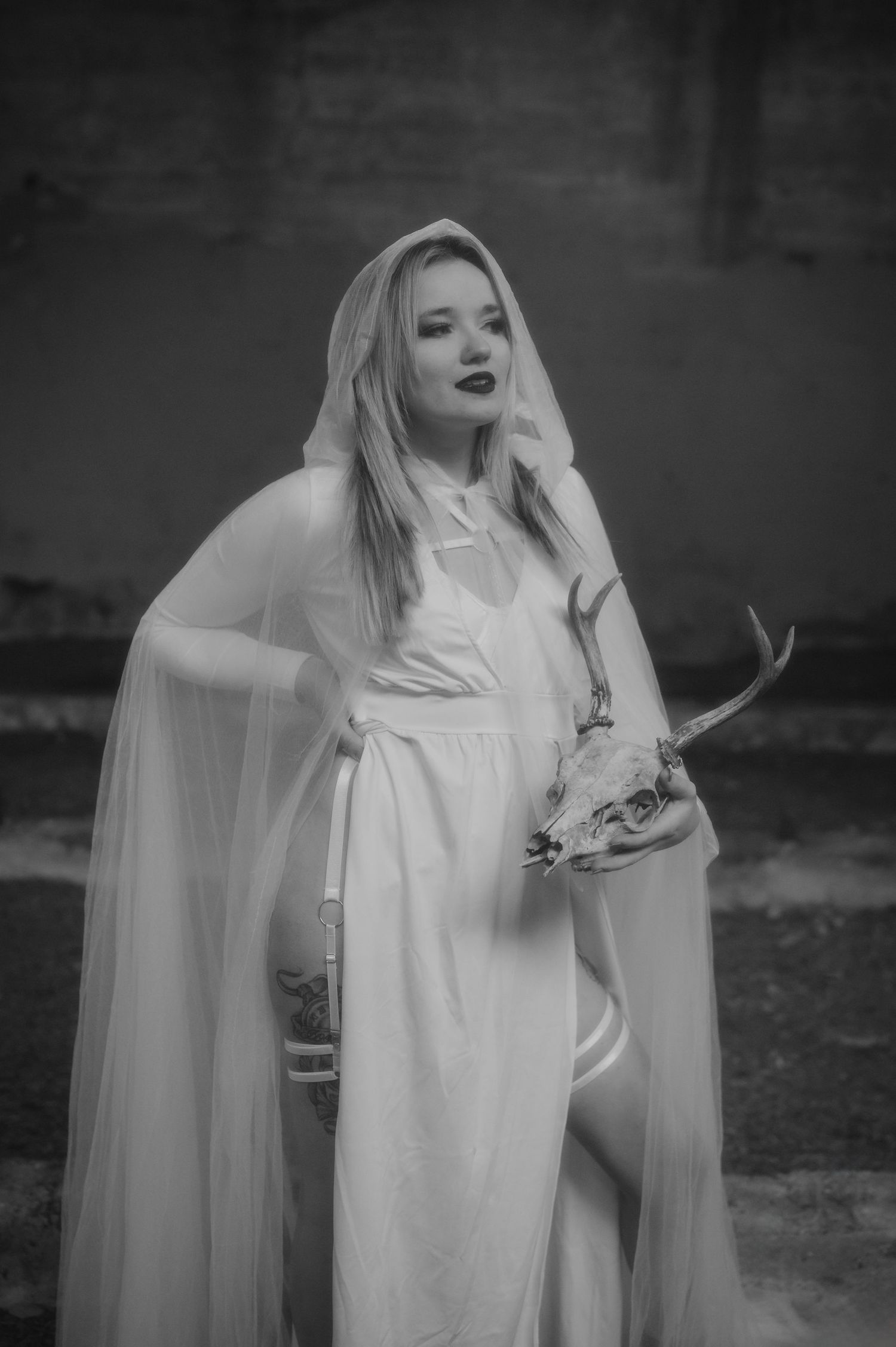 A figure in an ethereal white hooded cloak holds a deer skull in a moody black and white photograph.