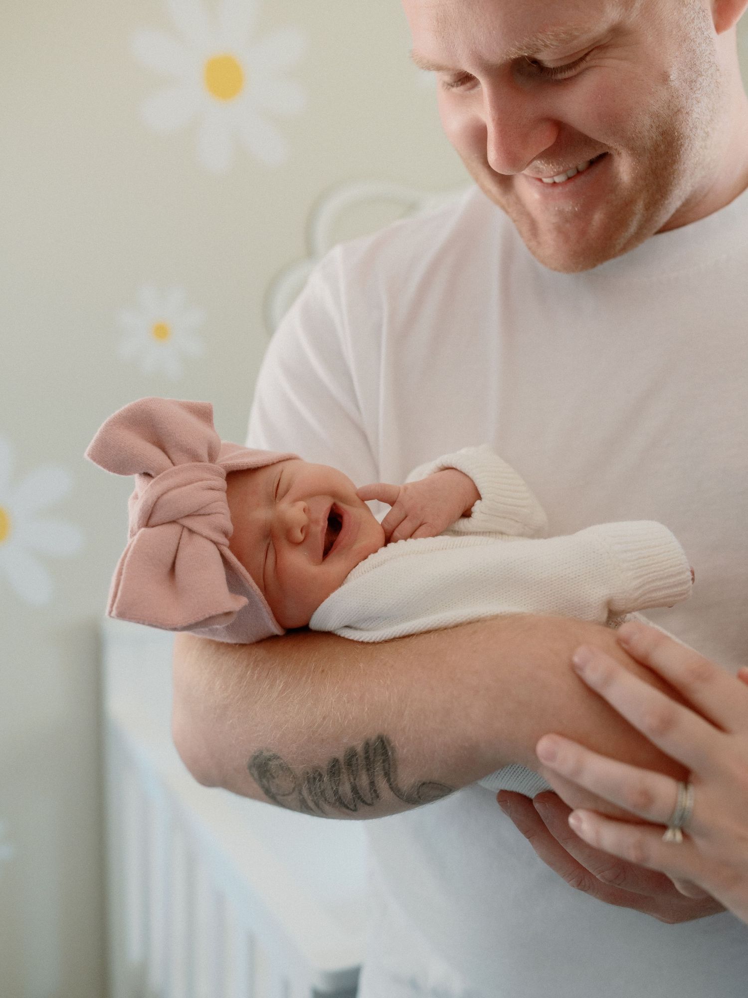 Parent in white shirt tenderly holds a yawning newborn baby wrapped in soft beige blanket against floral wallpaper background.