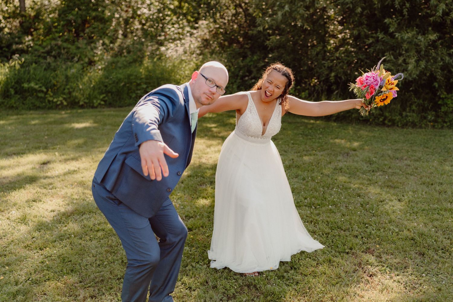 Playful wedding couple having fun while posing with colorful bouquet in a garden setting.